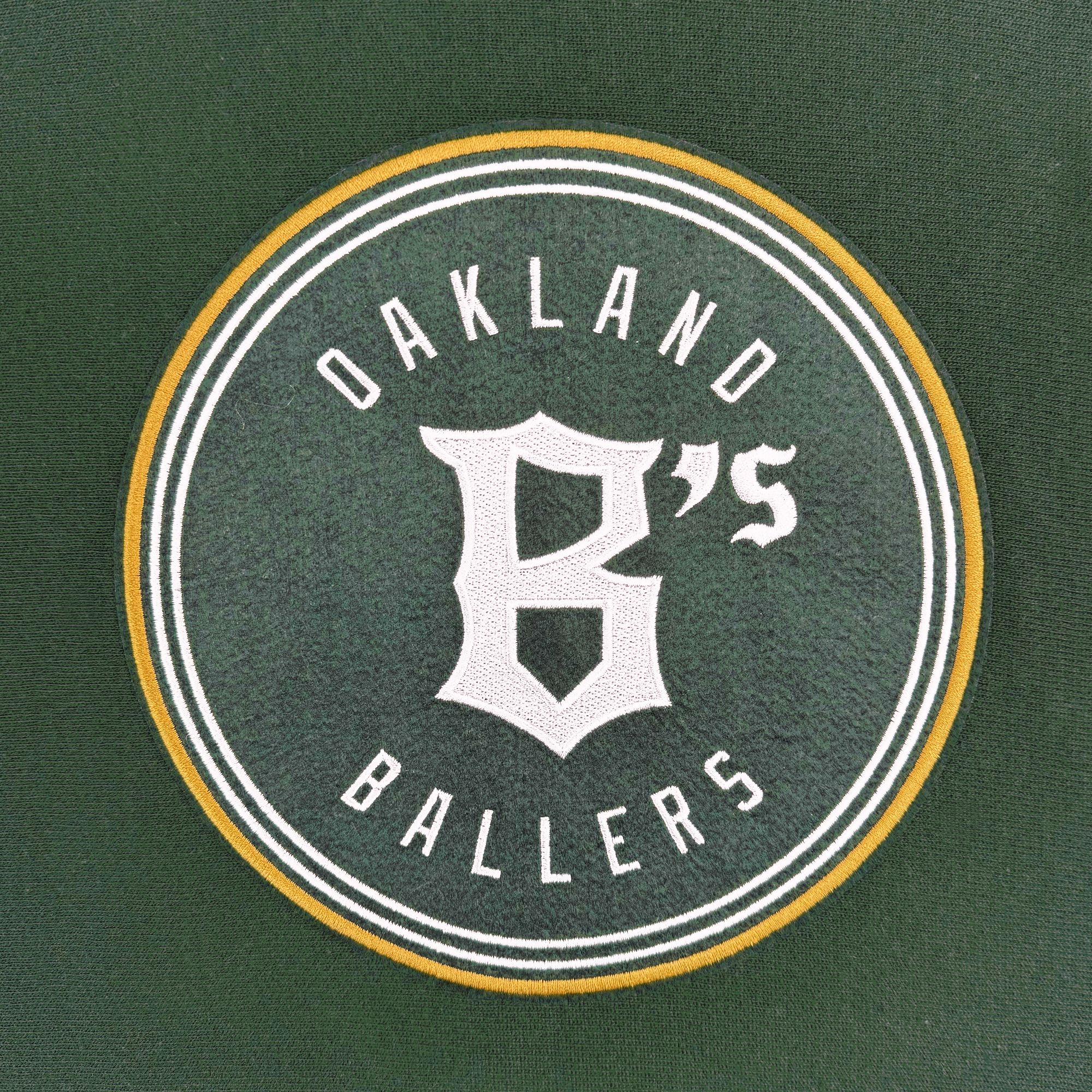 On a green background, the Oakland Ballers logo is circular with a yellow outline. Inside, "Oakland Ballers" is displayed in white text to celebrate the new pro baseball team's entry into the Pioneer League, and features a large, stylized "B" in the center. Get this design on the Champion X Oakland Ballers Logo Hoodie.