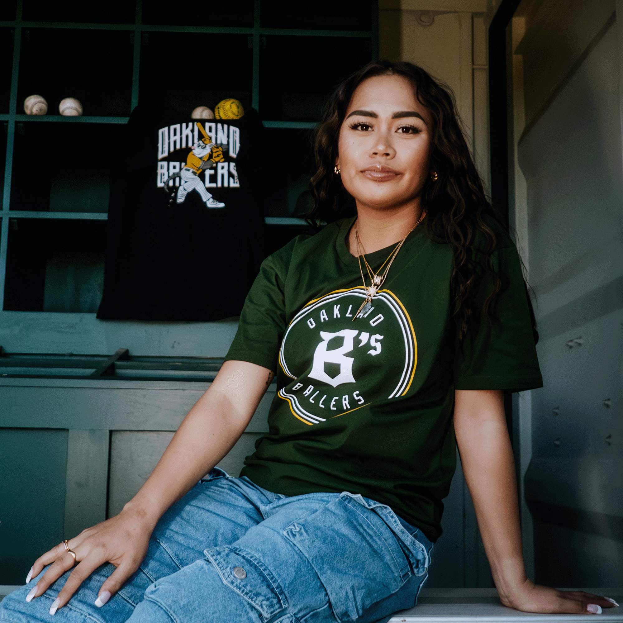 A woman relaxes on a wooden bench, sporting the Oakland Ballers Classic Tee in green paired with ripped jeans. Her wavy hair drapes under gold hoop earrings. A dark green shelf nearby displays baseballs and a black Oakland Ballers shirt, reflecting Pioneer League pride.