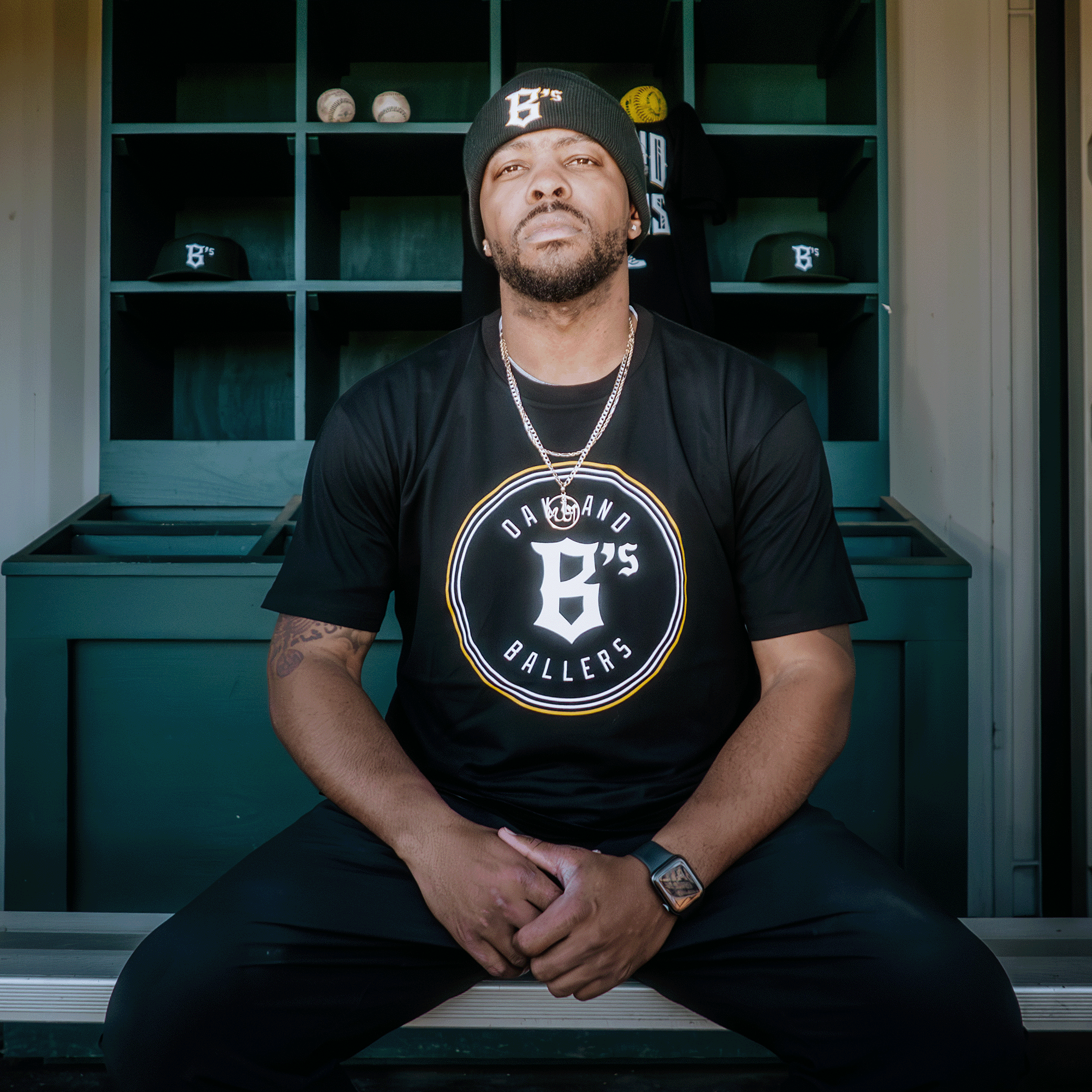 A man sits on a bench in front of a sports locker wearing an "Oakland Ballers Classic Tee" and matching beanie. Behind him, shelves are filled with baseball caps and balls. His serious look, beard, chain necklace, and watch highlight trademarks of the professional Pioneer League baseball team.