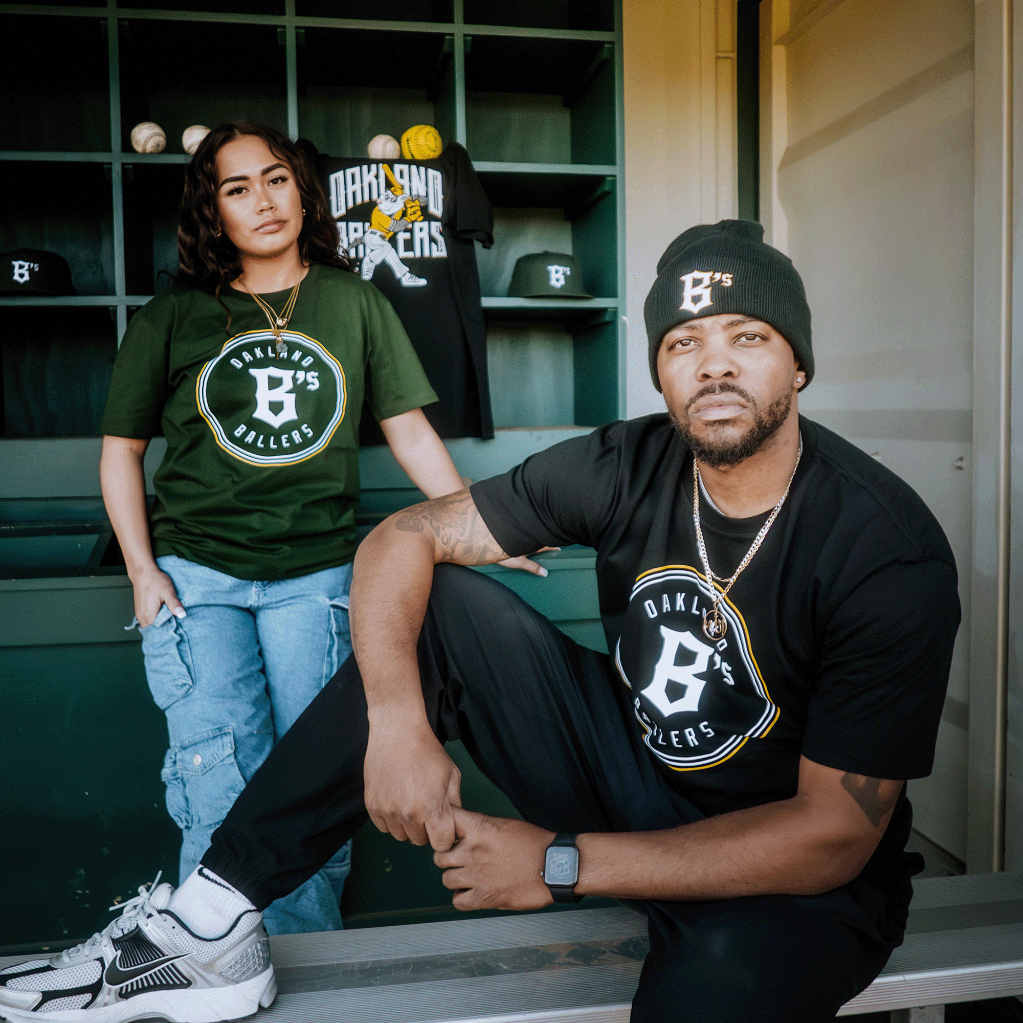 A man and woman pose proudly in front of green lockers. The man, seated, wears an "Oakland Ballers Classic Tee" and a beanie with sneakers, while the woman stands tall in a green Pioneer League shirt. Another "Oakland Ballers" tee hangs stylishly behind them.