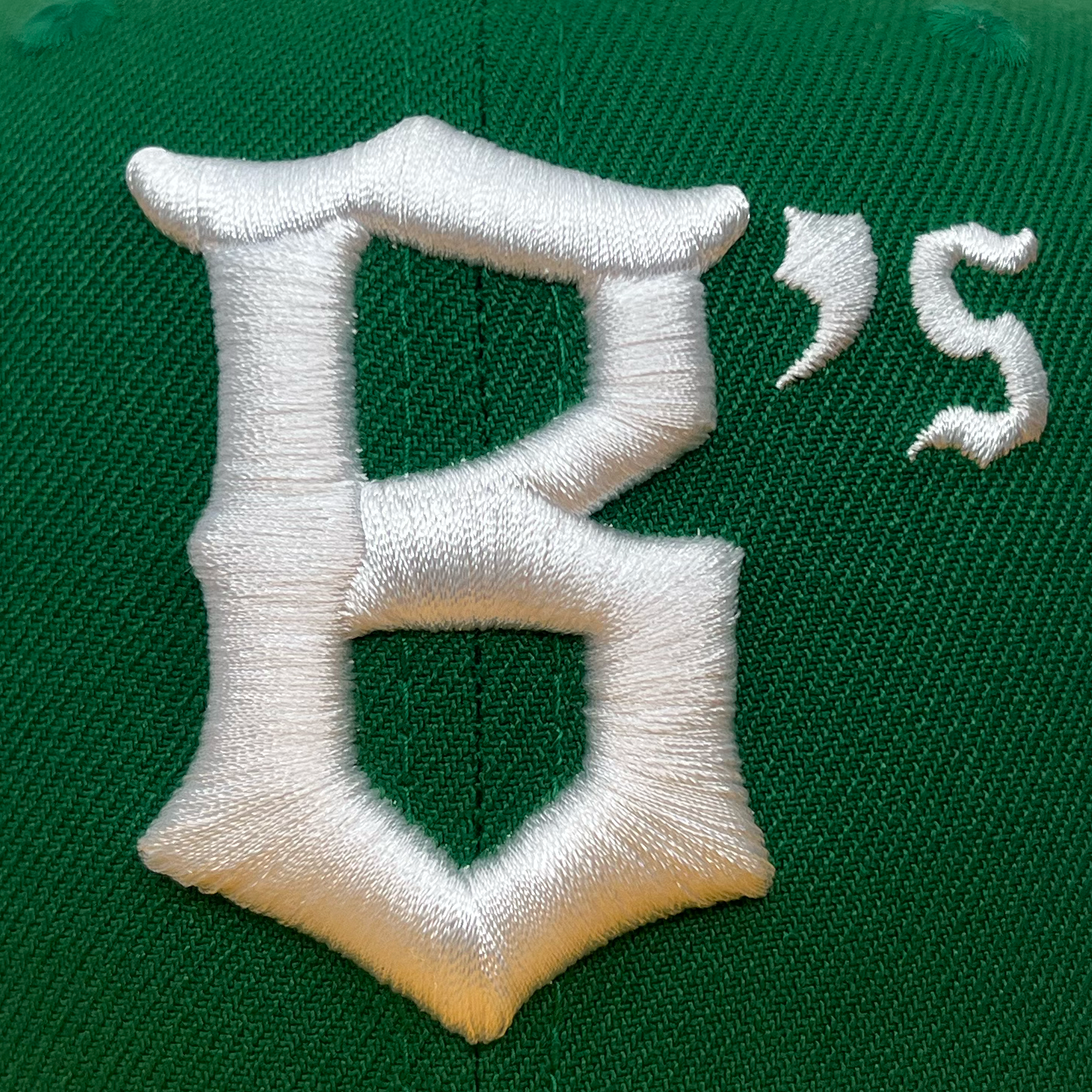 The New Era Oakland Ballers 59Fifty Cap is green and features a prominent, raised, white letter "B" embroidered on it. Next to the "B," in a slightly smaller font, is an apostrophe followed by a lowercase "s," also in white. The embroidery stands out against the green fabric of the cap, embodying the spirit of the Oakland Ballers.