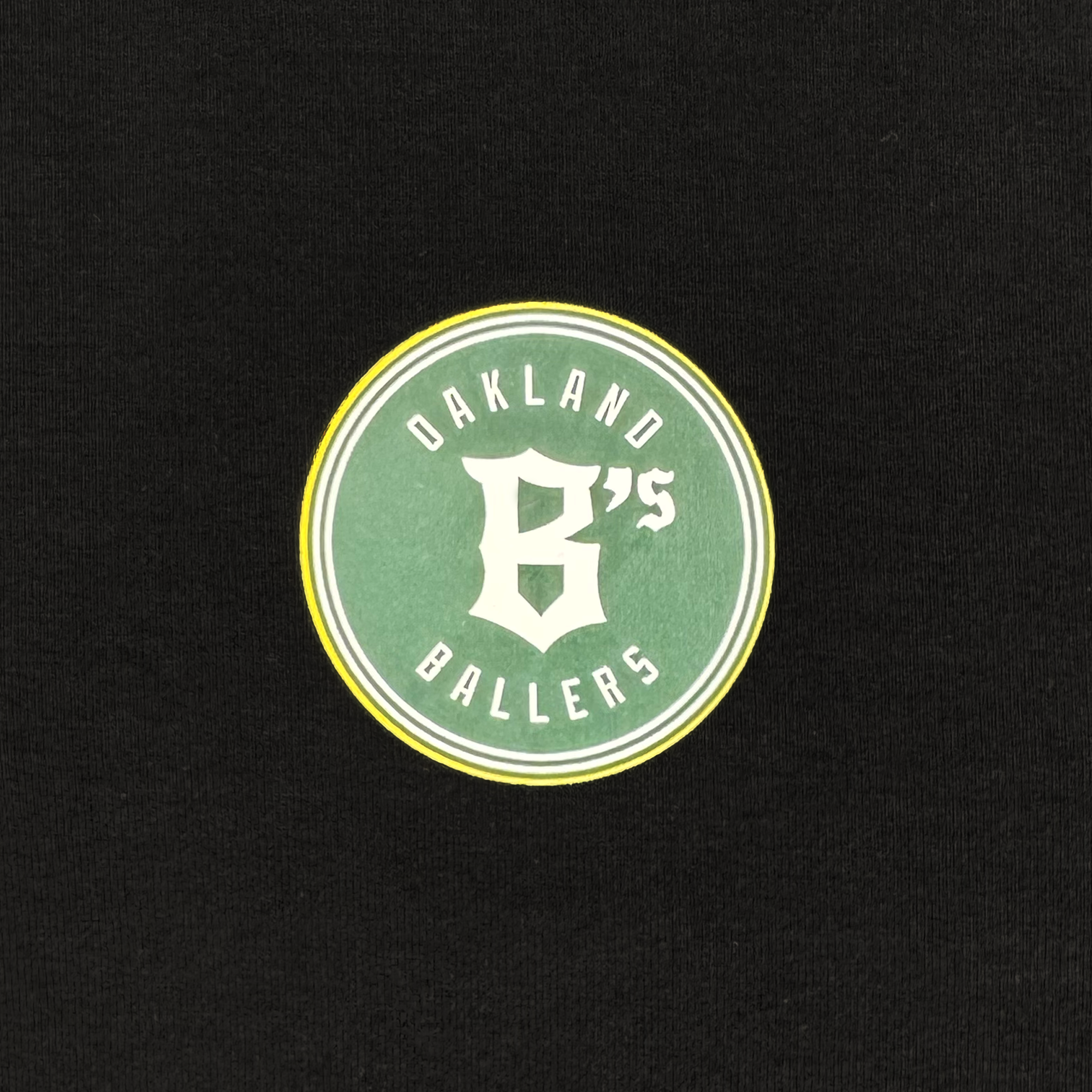 The Oakland Ballers team emblem is a round green patch with a yellow ring on black, featuring "Oakland Ballers" in white text. At the center is a stylized white "B's." It's the emblem of Oakland's Pioneer League baseball team, available on their Classic LS Hoodie Tee.