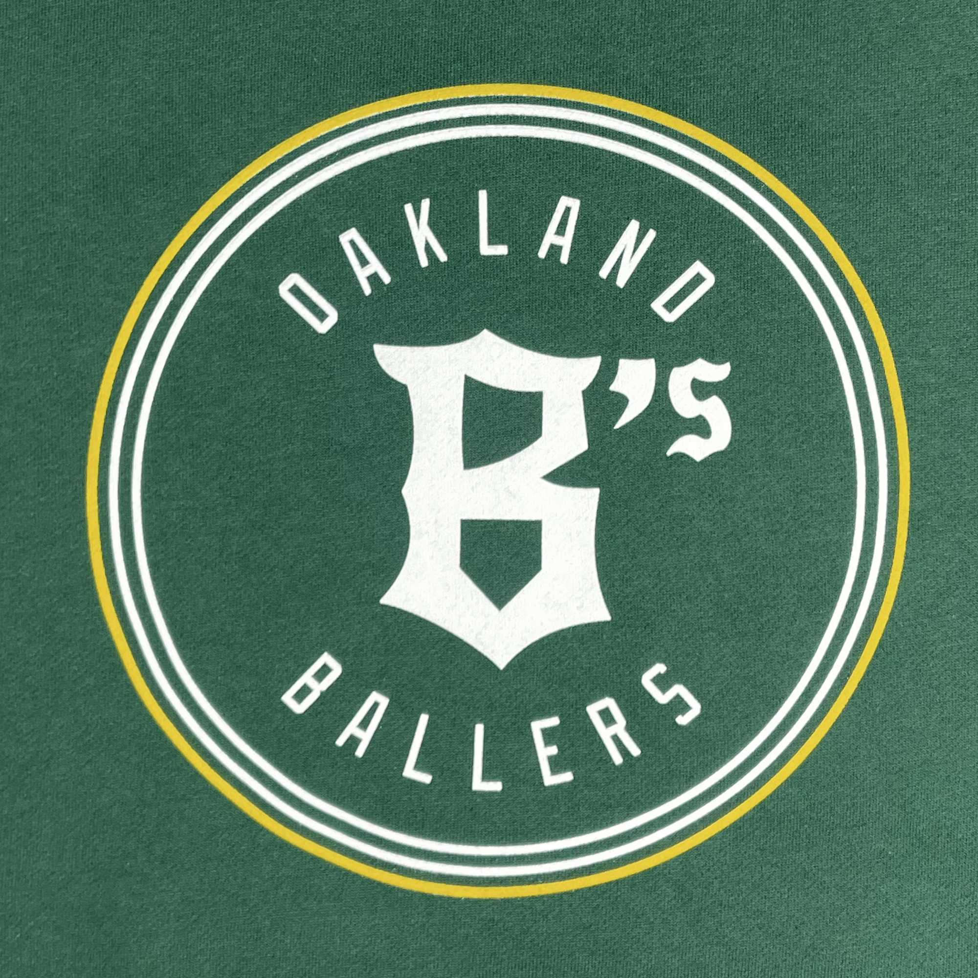 The Oakland Ballers Logo Heavyweight Hoodie highlights a circular logo on a green background with the text "OAKLAND BALLERS" in white, where "BALLERS" curving along the bottom. At the center, it showcases a stylized white letter 'B' followed by a small 's. Symbolizing Oakland's new pro baseball team in the Pioneer League, the logo is encircled by two white rings outlined by a thin yellow ring.