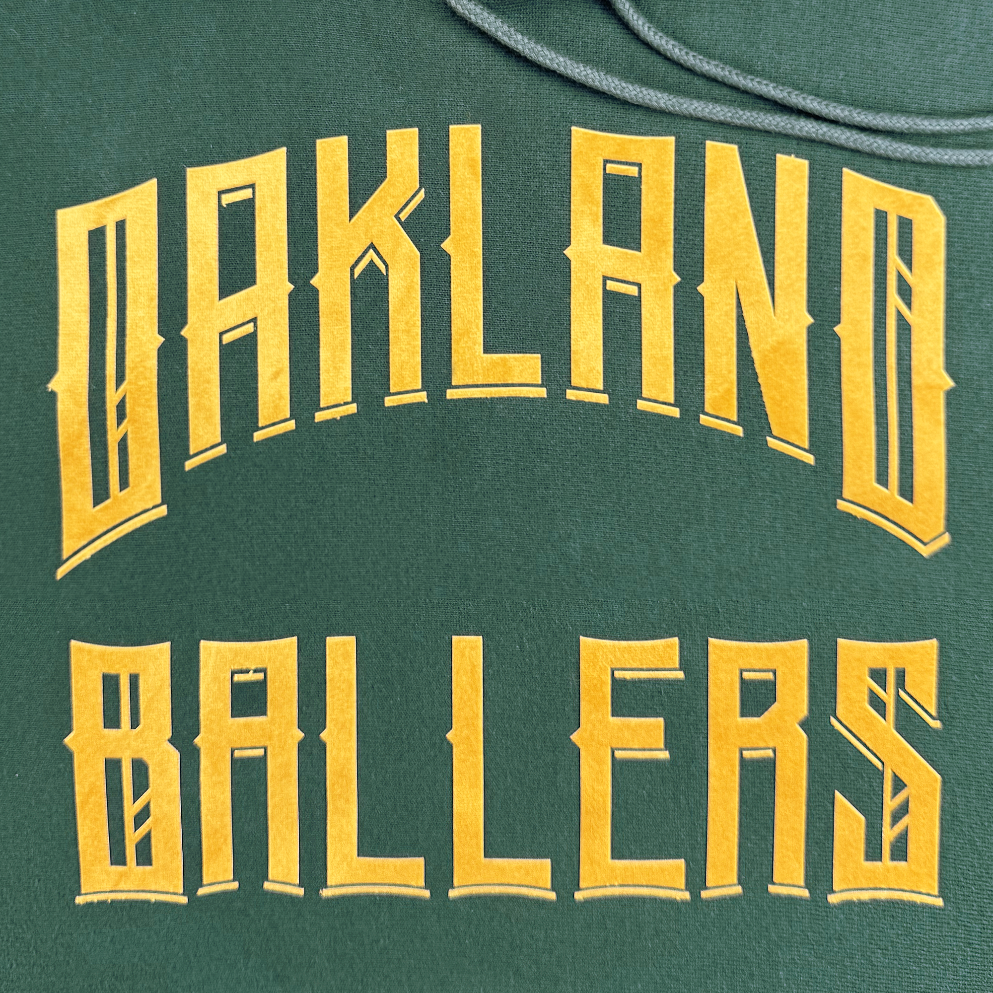 The Champion X Oakland Ballers Wordmark Hoodie showcases "Oakland Ballers" in large, bold yellow letters with a stylized angular font, ideal for fans of the Oakland baseball team in the Pioneer League. A hood drawstring is visible at the top edge of the image.