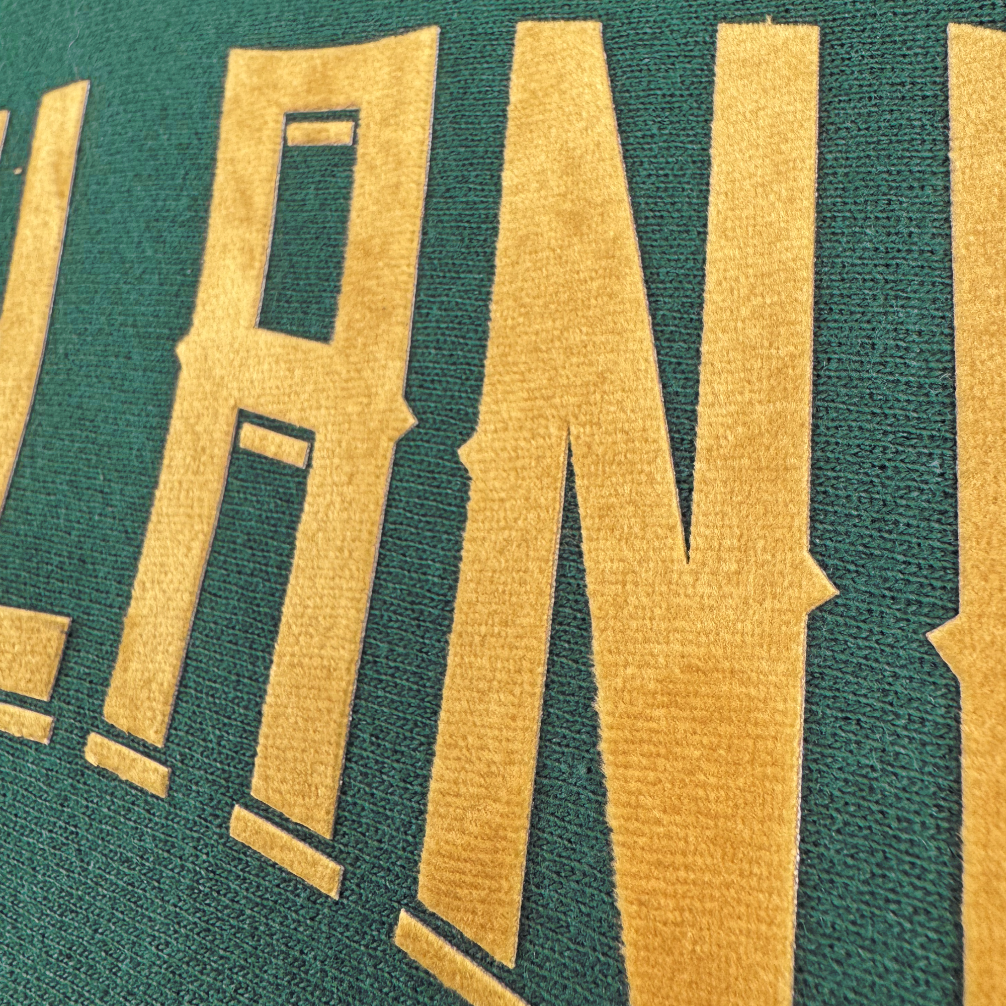 A close-up of the Champion X Oakland Ballers Wordmark Hoodie shows textured green fabric with bold, gold letters forming "LAND," inspired by Oakland Ballers' pride-filled jerseys. The texture adds depth, enhancing the standout contrast of the gold against green.