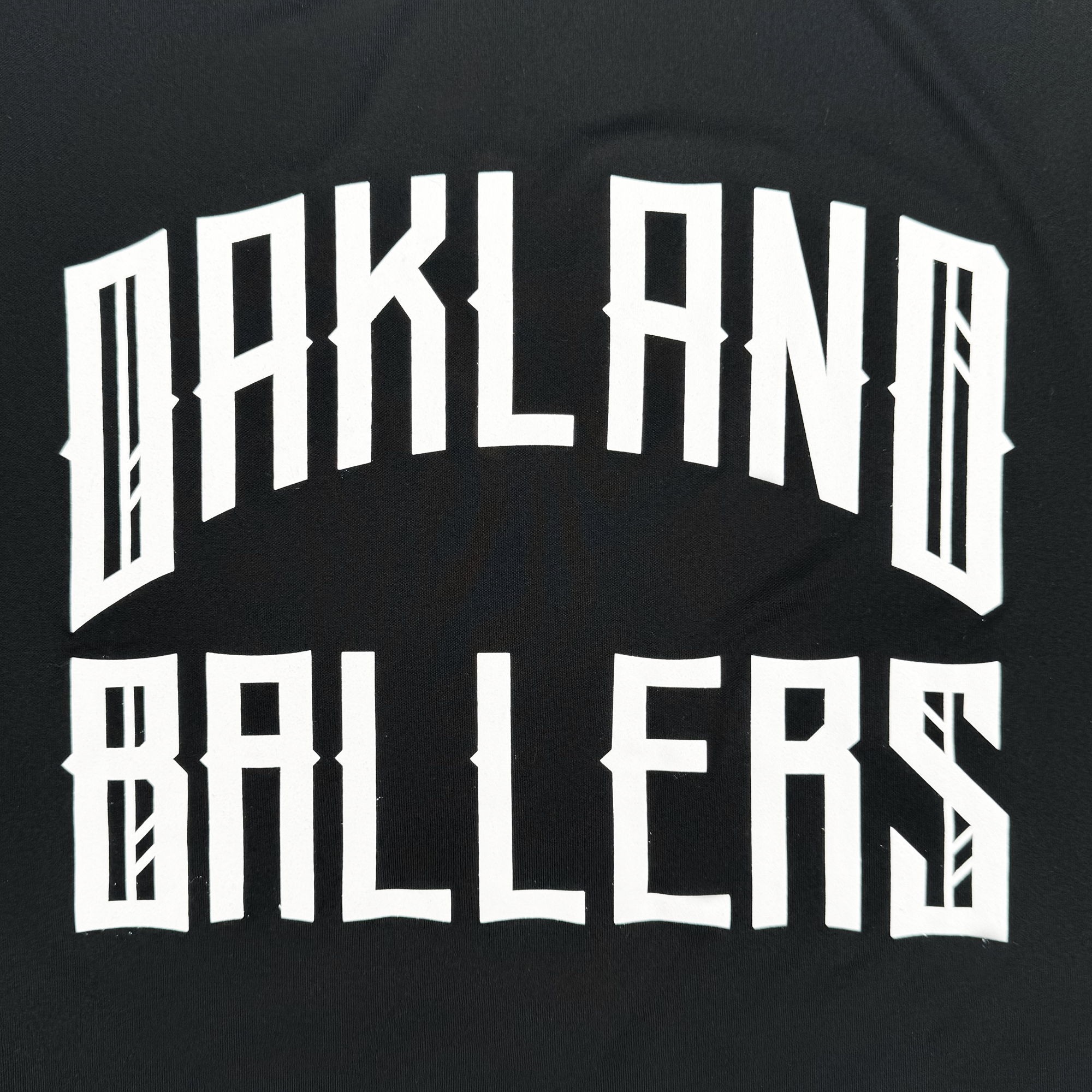 The image features "Oakland Ballers" in bold, white, stylized capital letters with a geometric design, subtly arched against a black background. This striking logo represents the new Oakland Ballers pro baseball team and appears on their Classic LS Hoodie Tee.