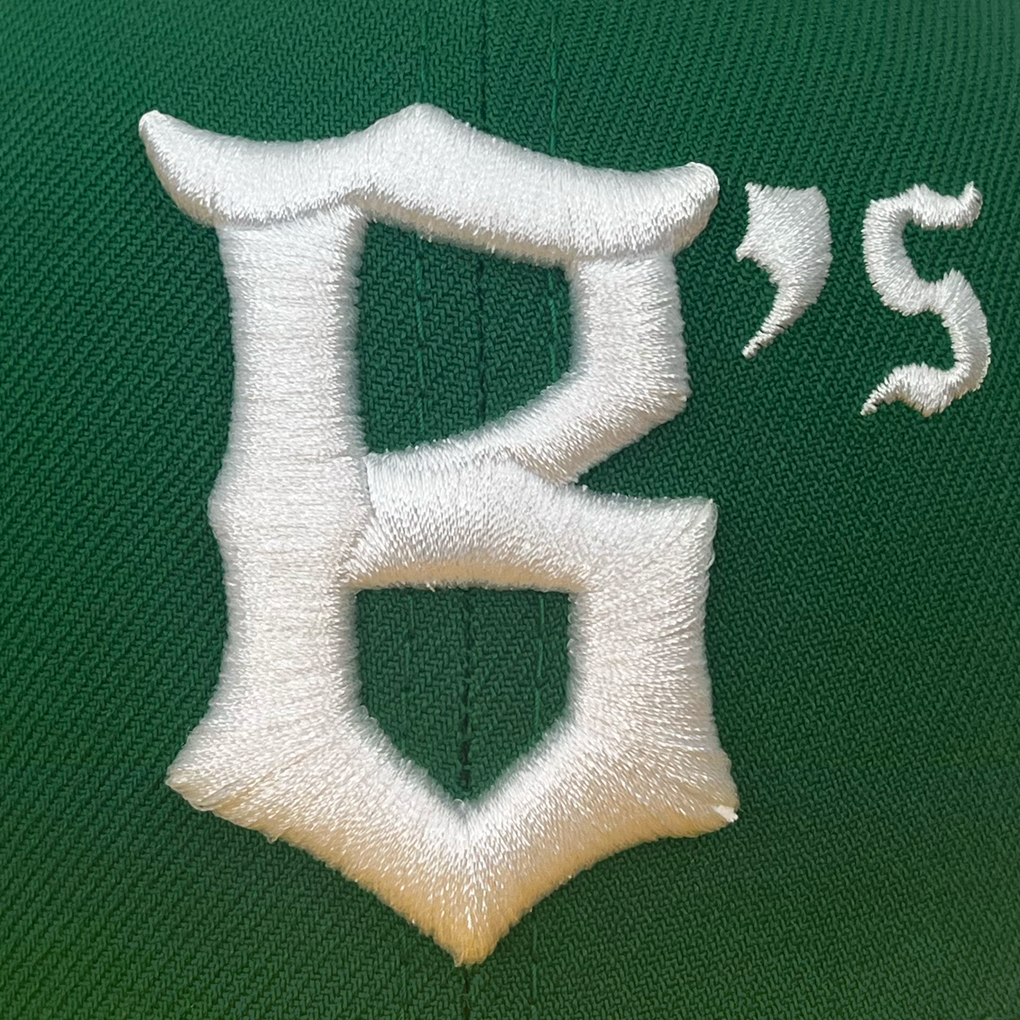 Close-up of a green fabric with a large, white, embroidered letter "B" and a smaller "'s" to its right, reminiscent of Pioneer League flair on the New Era Oakland Ballers 9FIFTY Cap. The stitching is detailed and prominent, giving a slightly three-dimensional texture to the text. The background is a solid green.