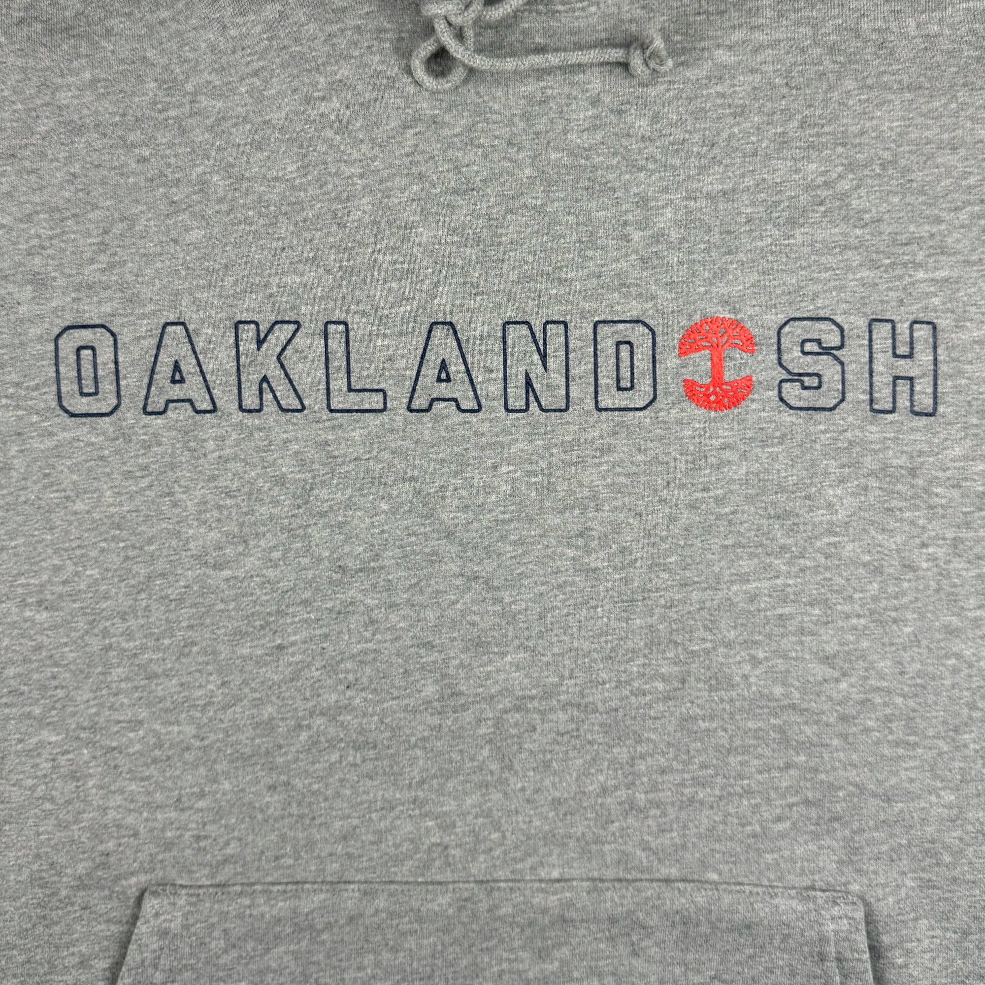 The Bandit Hoodie by Oaklandish is a heather grey pullover featuring "OAKLANDISH" in black letters across the chest, with a red tree ring design replacing the "I". Crafted from soft fabric, it includes drawstrings on the hood for a casual feel. Available in men's sizing for an ideal fit.