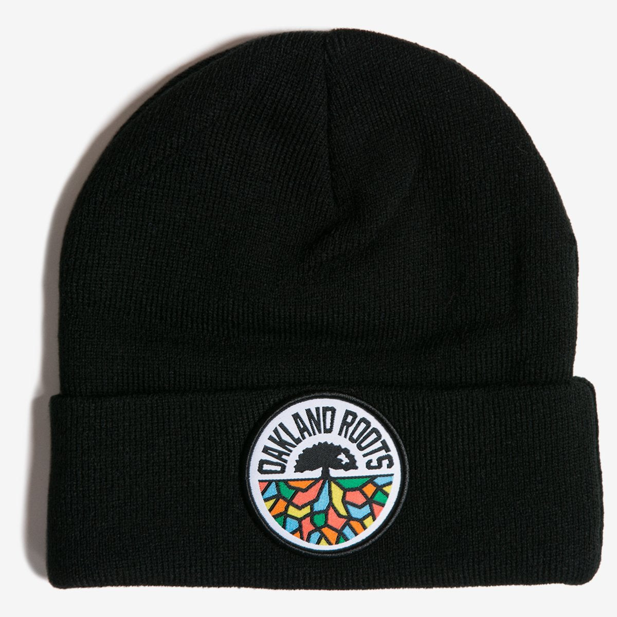 The Oakland Roots SC Cuff Beanie, crafted from black acrylic, showcases a folded cuff adorned with a circular patch. The patch prominently features a tree emblem at the top, beneath which vibrant stylized roots in shades of orange, green, and blue spread out. Curving along the top edge of the patch are the words "Oakland Roots Sports Club.