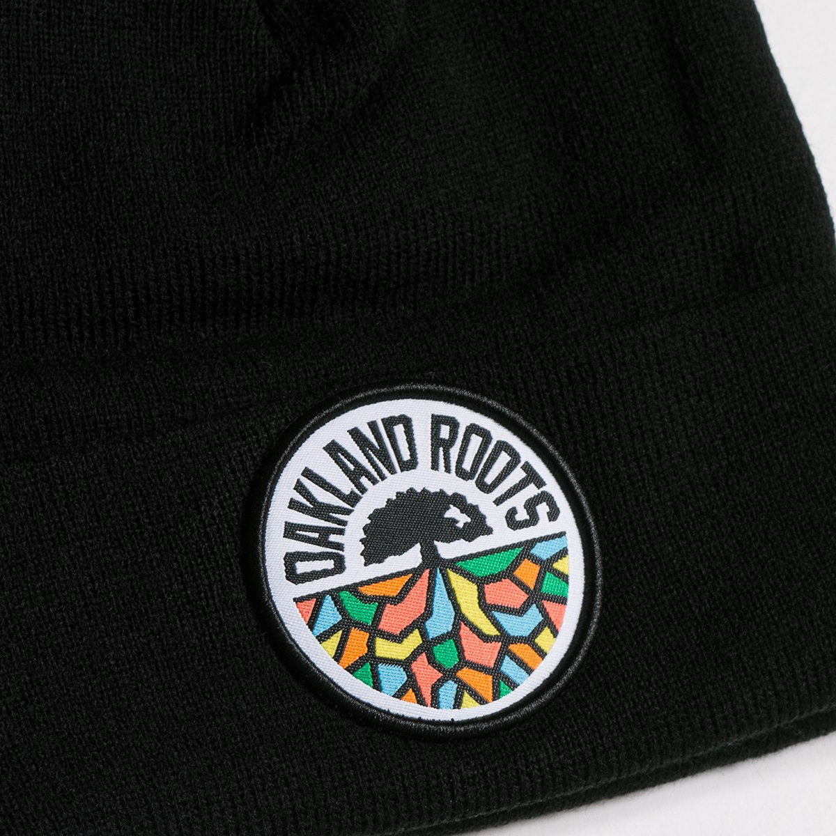 A close-up of the Oakland Roots SC Cuff Beanie by Oakland Roots SC, highlighting a circular patch. The patch proudly displays the text "OAKLAND ROOTS" around the top edge, with a black tree in the center and colorful abstract roots extending from the tree toward the bottom part of the circle in vibrant shades of orange, yellow, green, and red.