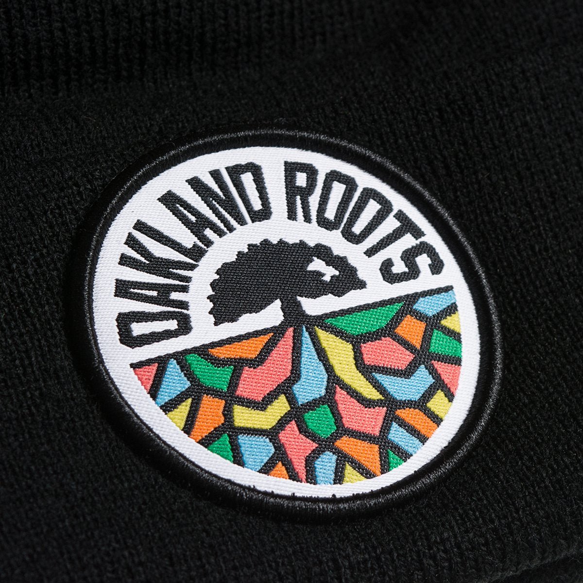 A detailed close-up of the Oakland Roots SC Cuff Beanie from the brand Oakland Roots SC. This black acrylic cuff beanie showcases a circular patch that reads "OAKLAND ROOTS" at the top. The patch features a silhouette of a tree with roots extending below, depicted in colorful geometric shapes in red, yellow, green, and blue.