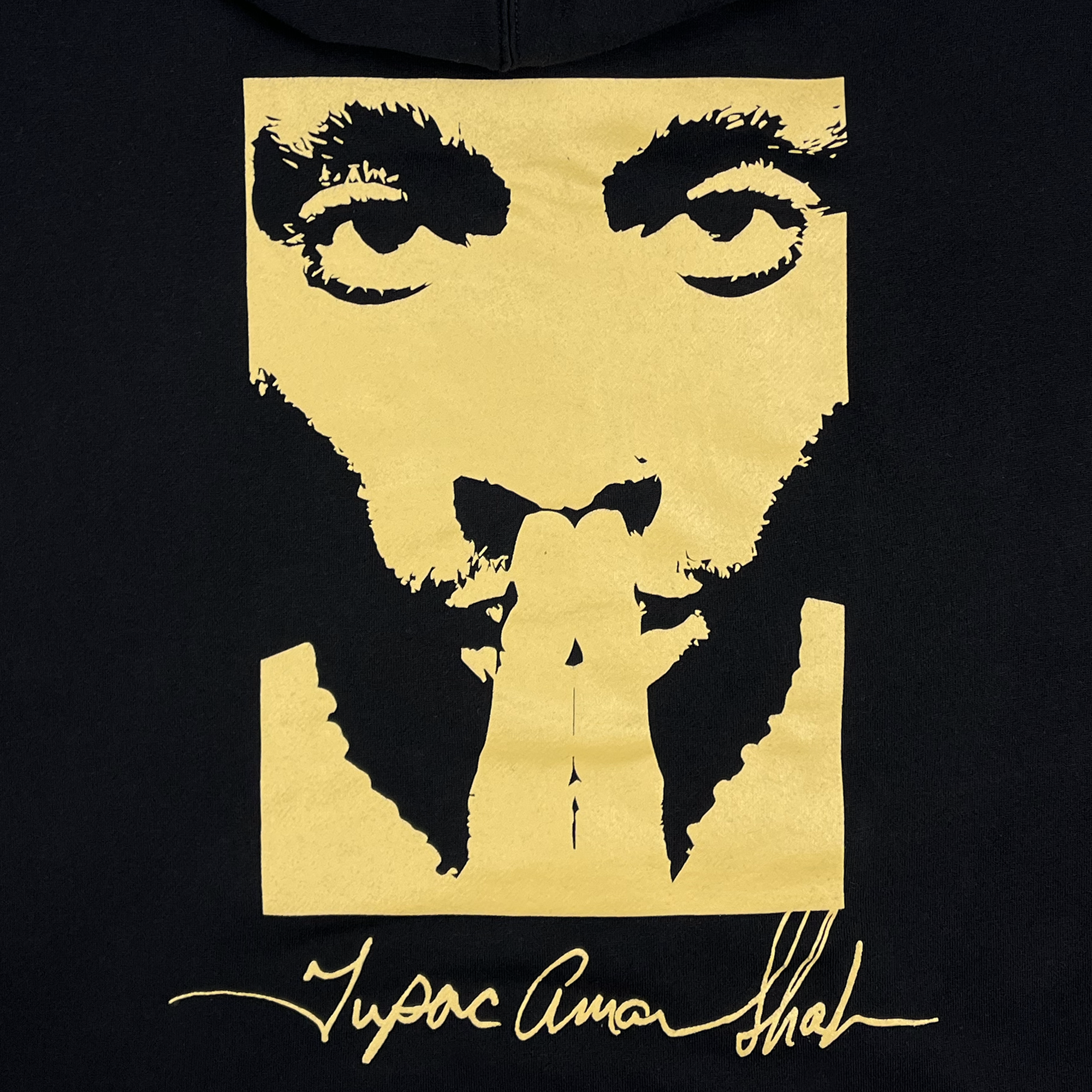 The Bless Hoodie from Oaklandish showcases a striking black background with a stylized golden portrait of a man gazing forward, hands clasped in a prayer-like pose, and features the elegant cursive signature "Tupac Amaru Shakur," celebrating his enduring legacy.