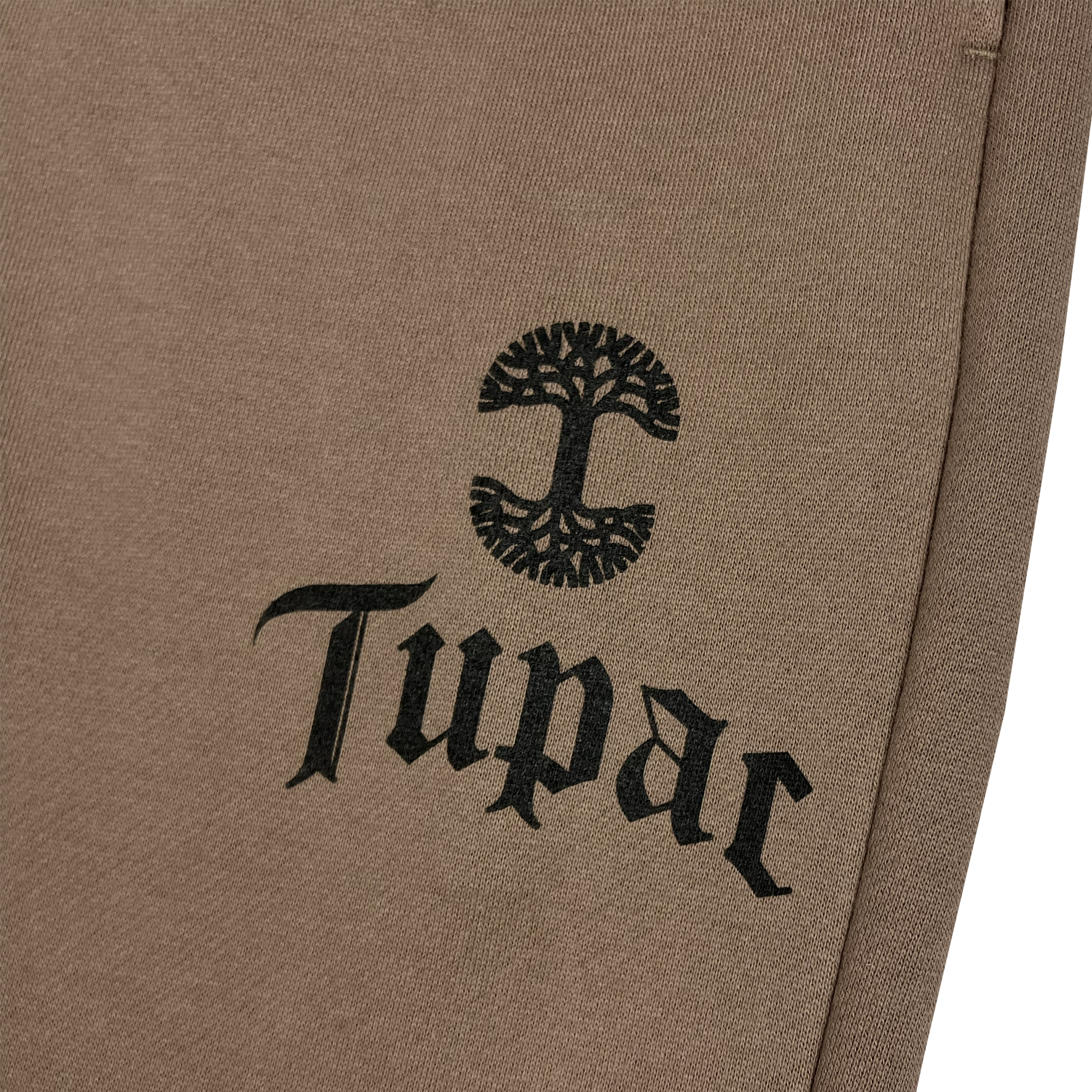 A detailed view of the Bless Jogger from Oaklandish shows beige fabric adorned with a black stylized tree emblem above the name "Tupac" in a gothic font. The textured material implies it's part of apparel, with the tree's intricate branches creating a circular design, suggesting a Tupac collaboration.