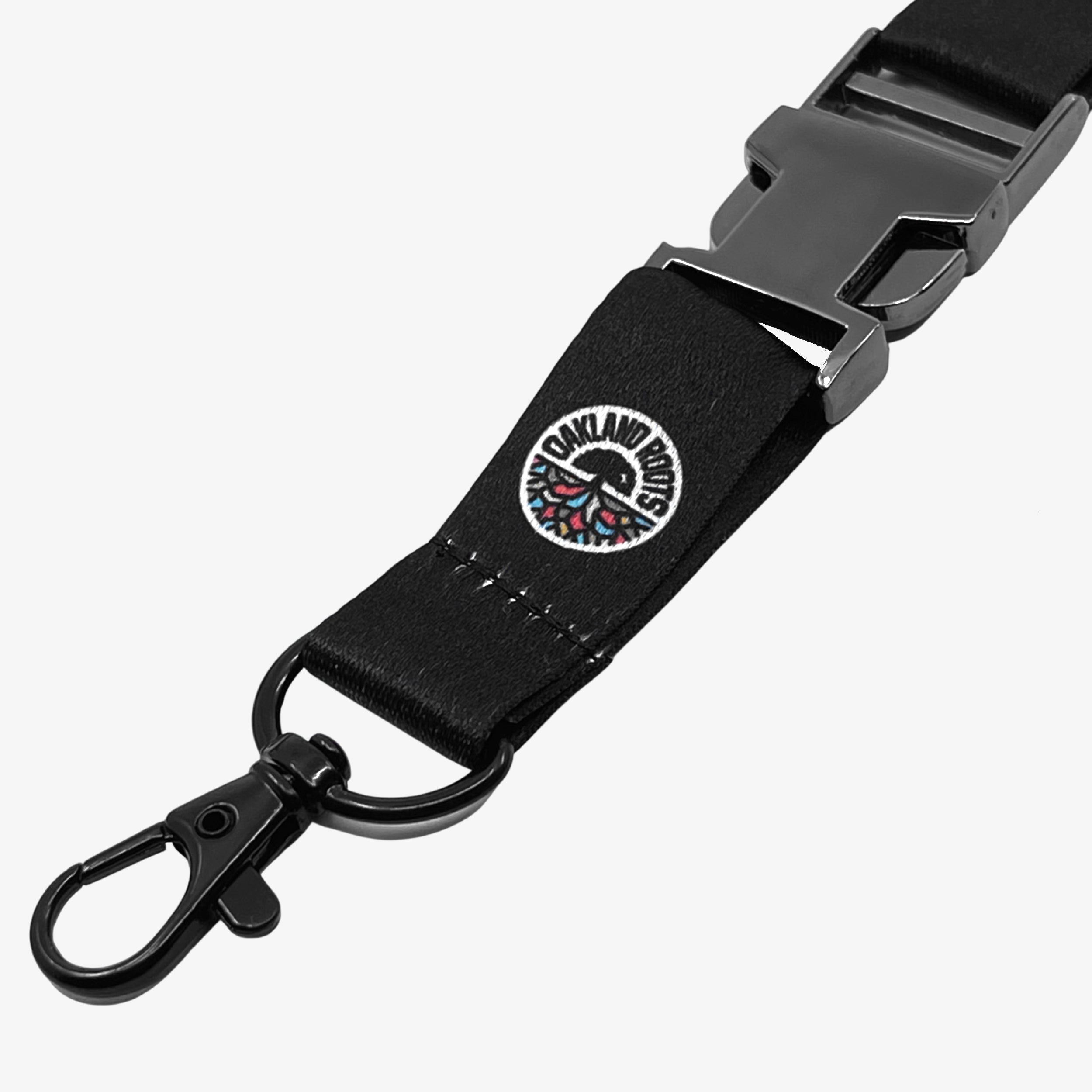 The Oakland Roots SC VIP Lanyard from Oakland Roots SC is black and features a metal clasp at one end, along with a circular embroidered patch near the clasp. The patch showcases a design with text reading "OAKLAND ROOTS SPORTS CLUB," celebrating the spirit of social good. Additionally, the lanyard includes a detachable plastic buckle.