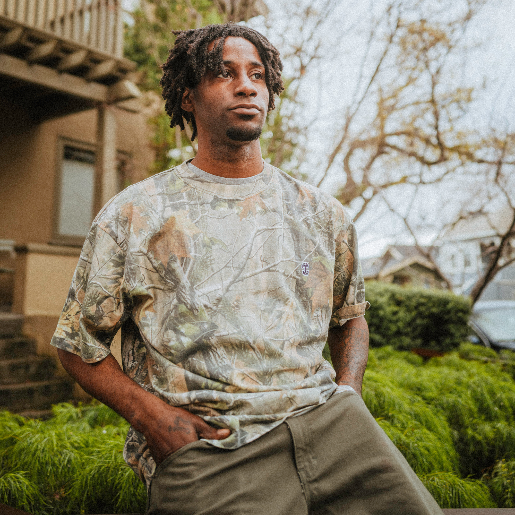 Wearing an oversized Oaklandish Camo Tree Tee and olive green pants, a person with short dreadlocks stands outdoors. Their camo-print shirt blends seamlessly with the peaceful tree camo backdrop under an overcast sky, creating a scene of deep contemplation.