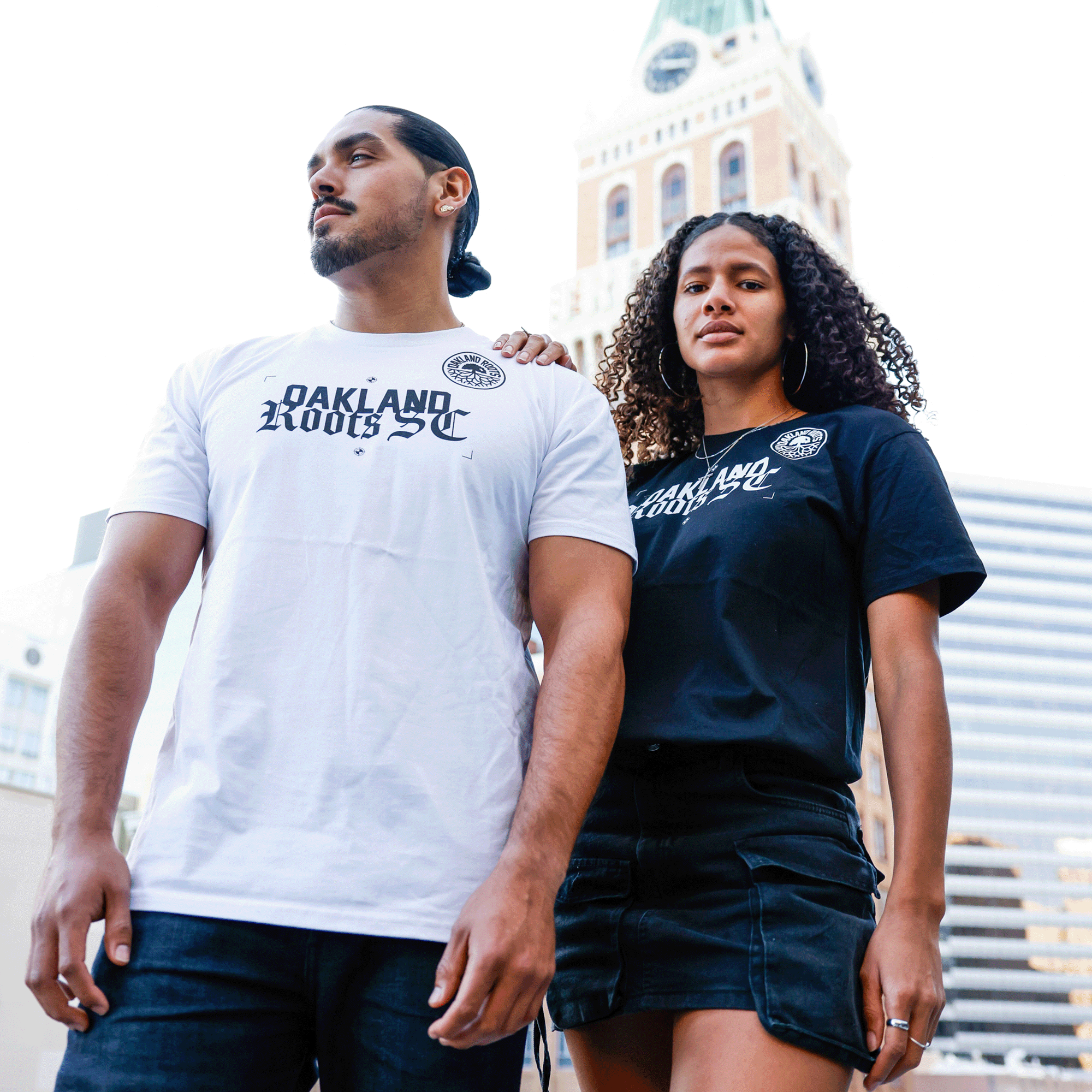 Two people stand outside in front of a tall building with a clock tower. The person on the left wears an "Oakland Roots SC Futbol Town Tee" from Oakland Roots SC and jeans, and has long hair tied back. The person on the right, wearing a black "Oakland Roots SC Futbol Town Tee" from Oakland Roots SC and a skirt, has curly hair.