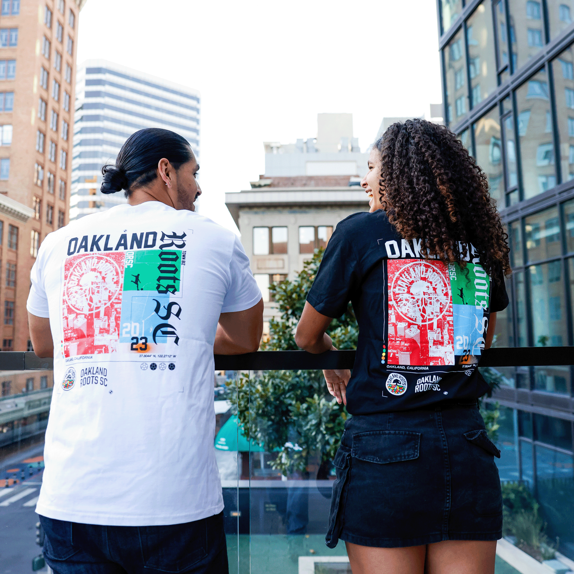 A man and a woman are standing on a balcony, seen from behind, both wearing Oakland Roots SC Futbol Town Tees. The man sports a white tee, while the woman opts for black. They appear to be smiling at each other as they take in the view of tall buildings and trees in Oakland, proudly supporting their sports club.