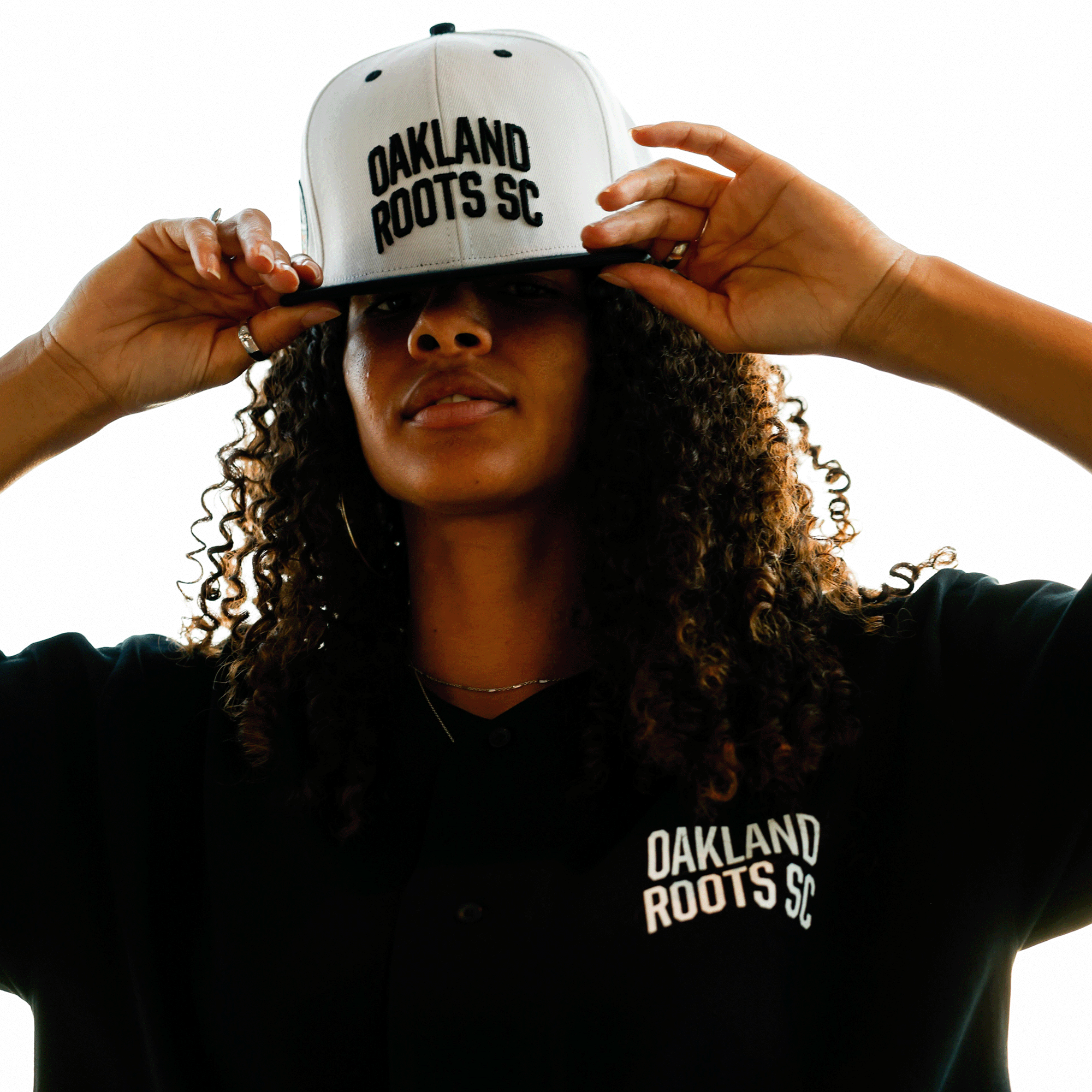 A person with curly hair wears a black shirt with "Oakland Roots SC" on the chest and adjusts an Oakland Roots SC Arched Text Snapback. Their face is partially shadowed, while the bright white background emphasizes their silhouette and the clothing's logo.