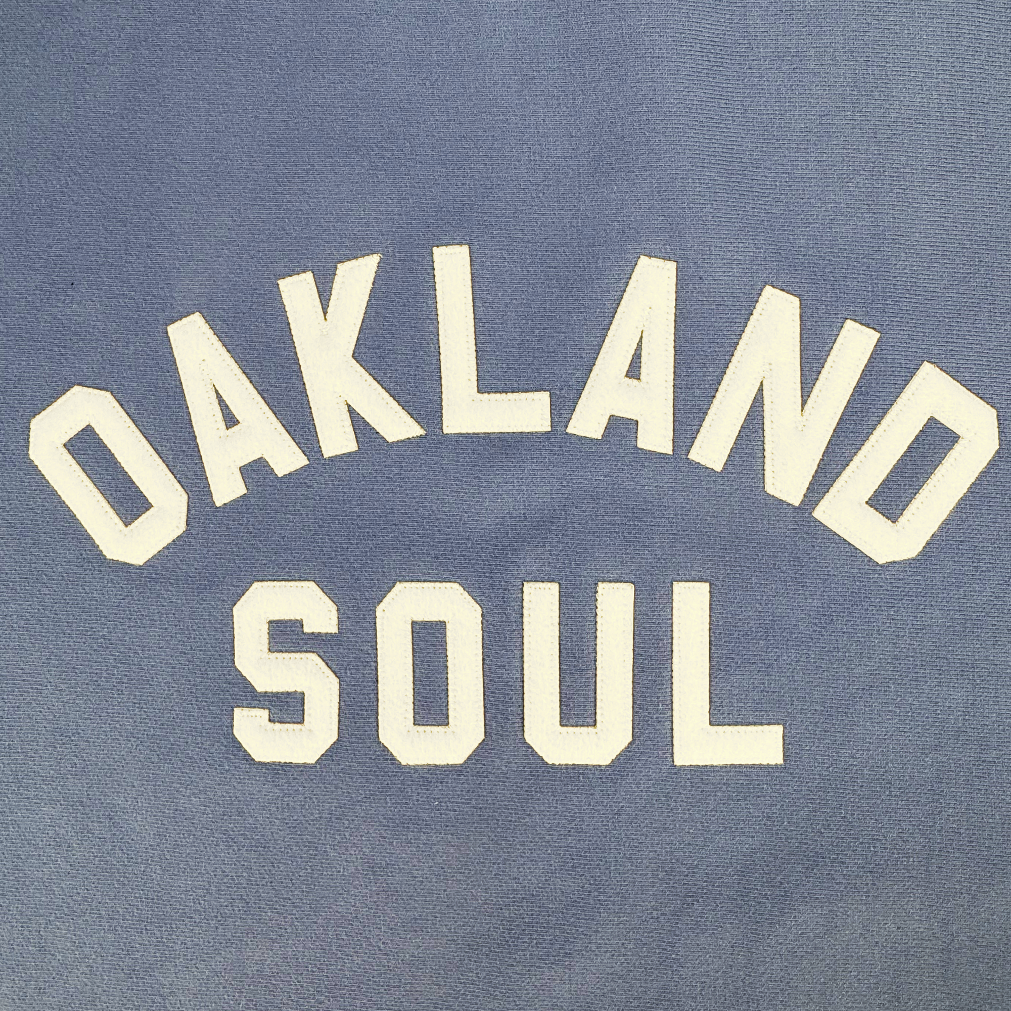 The Champion X Oakland Soul Wordmark Crew features blue fabric with bold uppercase white text. "OAKLAND" arches over "SOUL," celebrating the spirited play of Oakland Soul in the USL W-League, and is printed or embroidered to represent women's soccer.