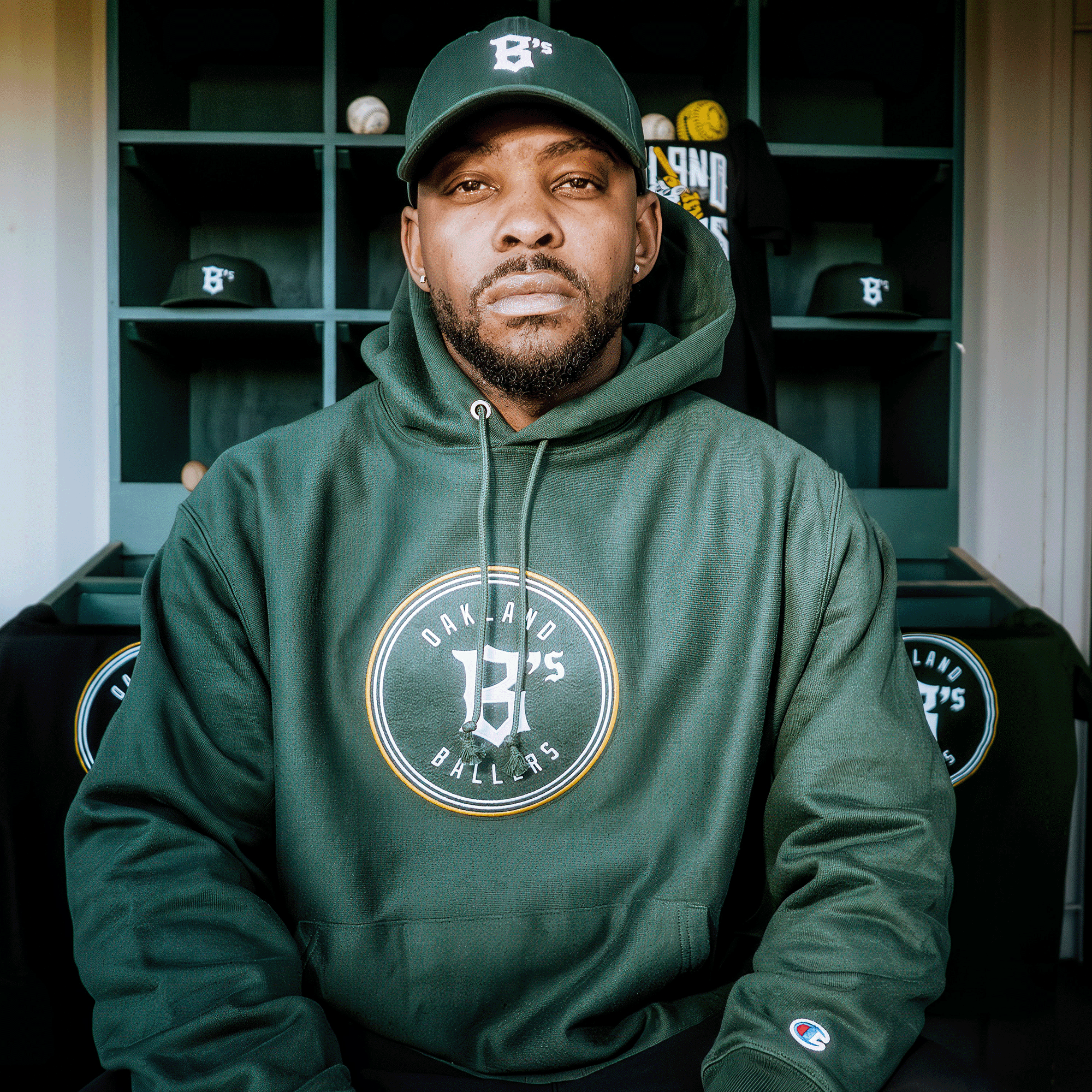 A man in a Champion X Oakland Ballers Logo Hoodie and matching cap, featuring Oakland's new pro baseball team logo, sits against a locker room backdrop. His serious expression and nearby lockers filled with caps and sports gear reflect the team's ambitions in the Pioneer League.