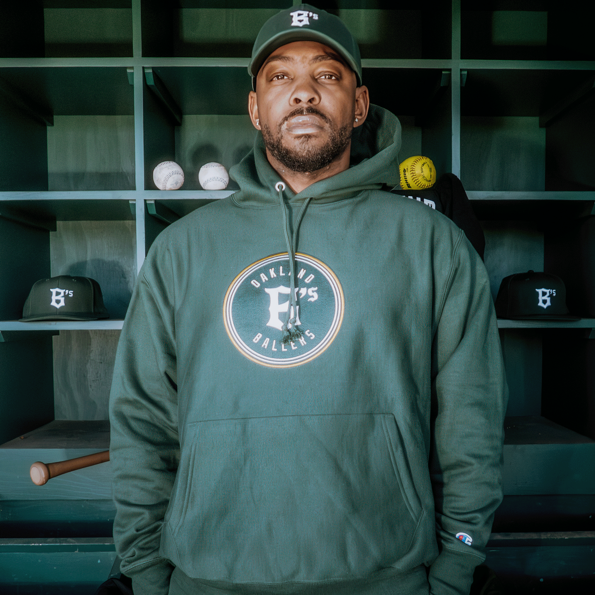 A man in a green Champion X Oakland Ballers Logo Hoodie and matching cap stands in a professional-looking sports dugout. Shelves behind him hold baseballs, caps, and a bat, hinting at his connection with the Oakland Ballers in the vibrant Pioneer League setting.