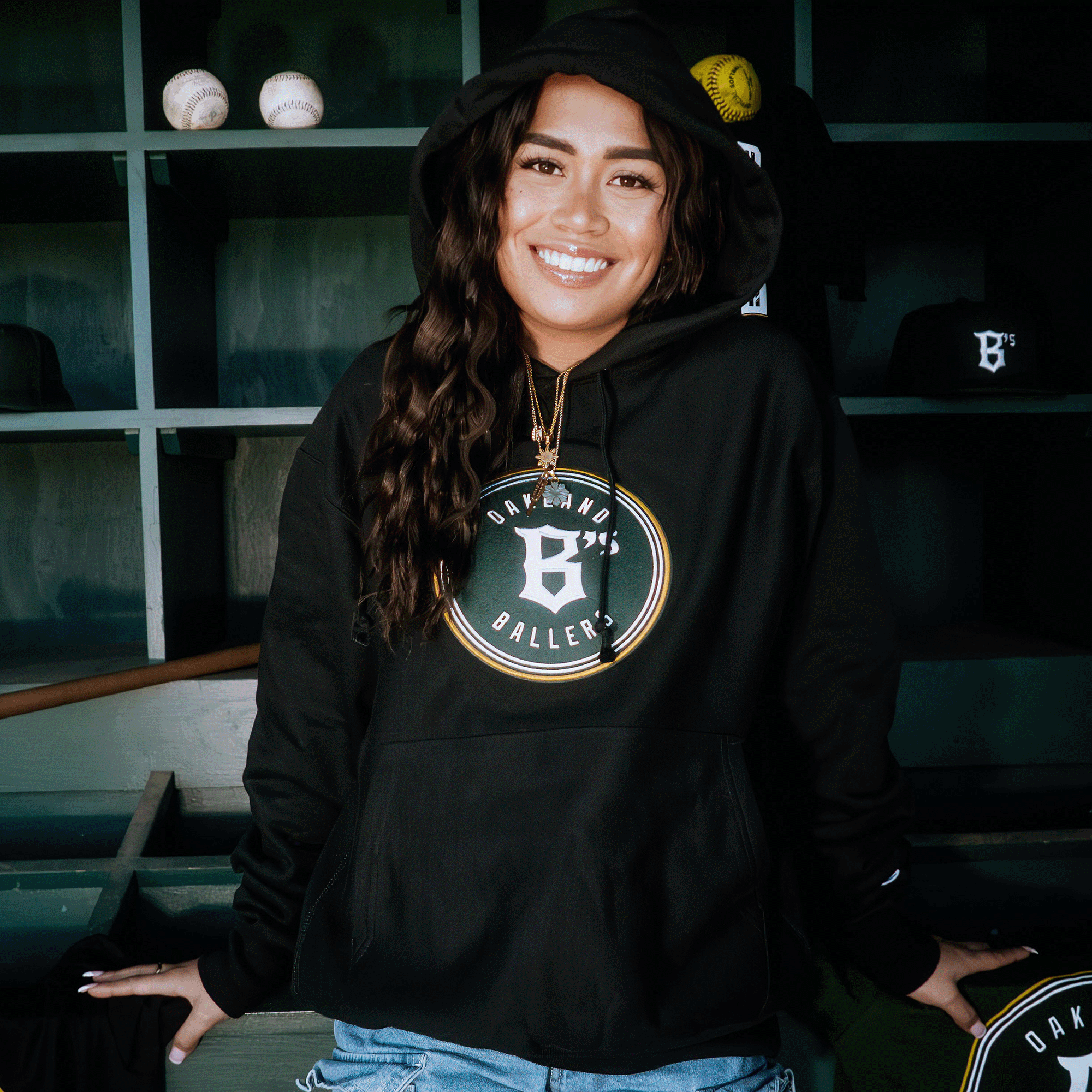 A cheerful person with long, wavy hair stands in front of a sports-themed backdrop wearing a Champion X Oakland Ballers Logo Hoodie. The backdrop is adorned with shelves of baseball caps and balls, celebrating their favorite Pioneer League team, the Oakland Ballers.