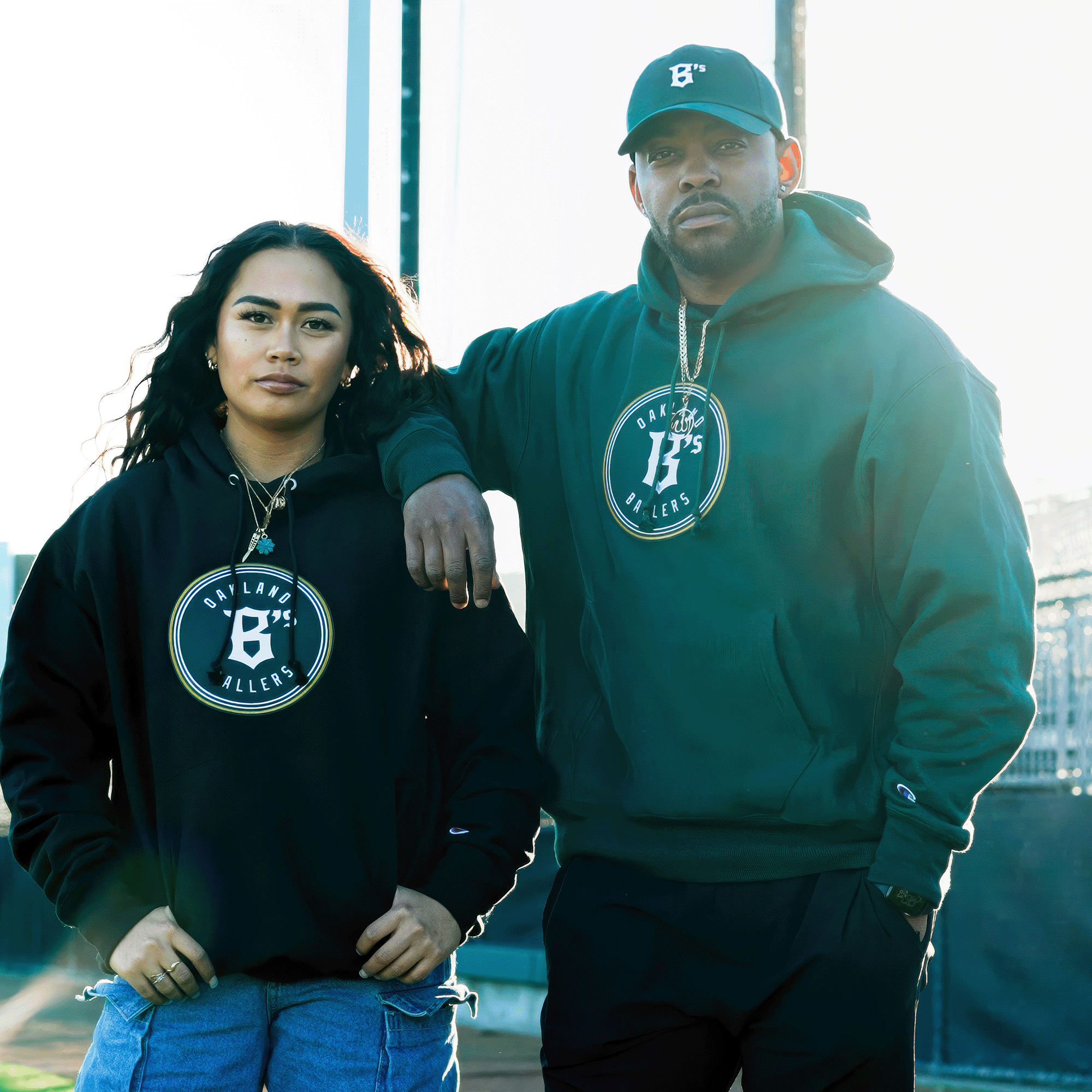 A man and woman pose outdoors in matching Champion X Oakland Ballers Logo Hoodies, with the man wearing a cap also featuring Oakland Ballers. The woman, with long hair and denim pants, stands confidently while he rests his arm on her shoulder against a sunlit city backdrop.