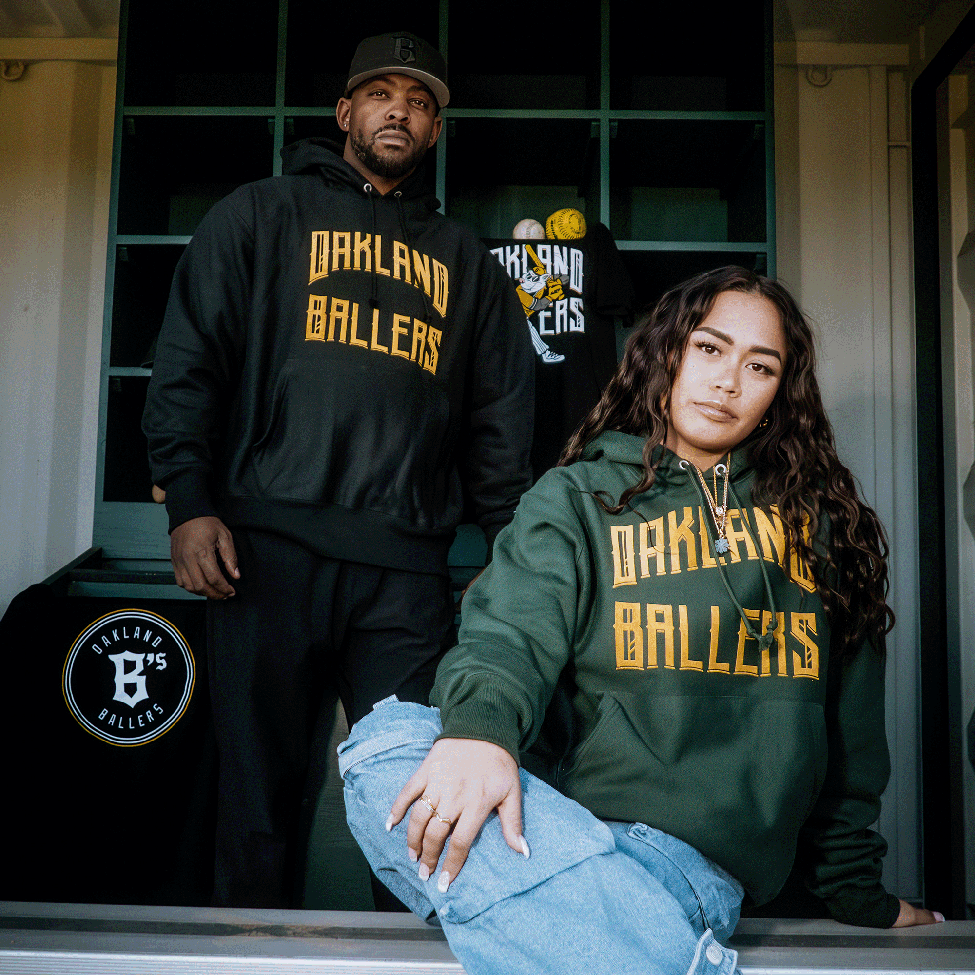 Two people wearing Champion X Oakland Ballers Wordmark Hoodies confidently represent Oakland's new pro baseball team. The left wears a black hoodie and the right a green one, with caps and T-shirts in the background. A circular "Oakland Ballers" logo is visible on a window.