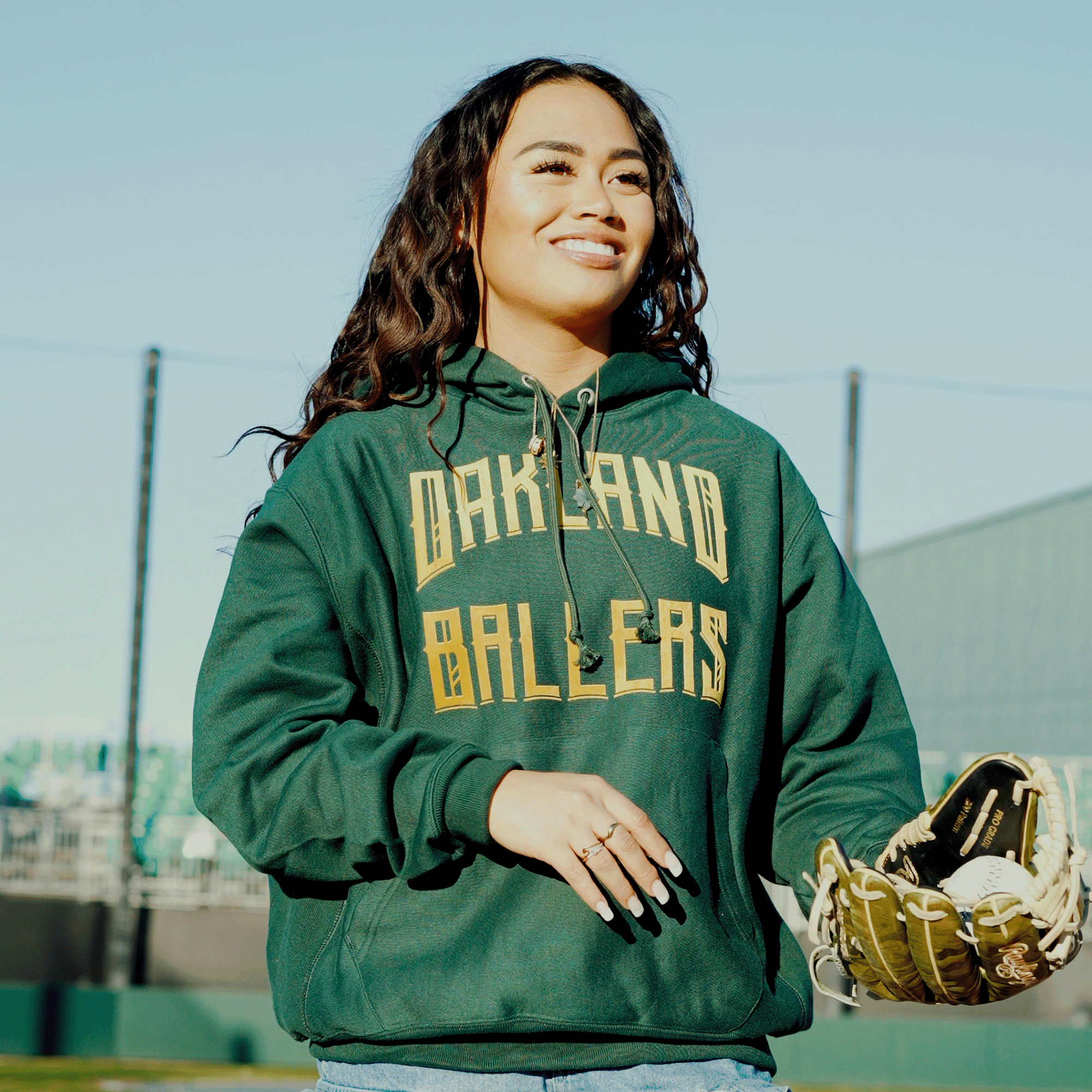 A long-haired person with a smile walks on a baseball field under clear skies, wearing the Champion X Oakland Ballers Wordmark Hoodie. The green hoodie features "Oakland Ballers" in gold letters. They’re holding a baseball glove, and the field is fenced in the background.