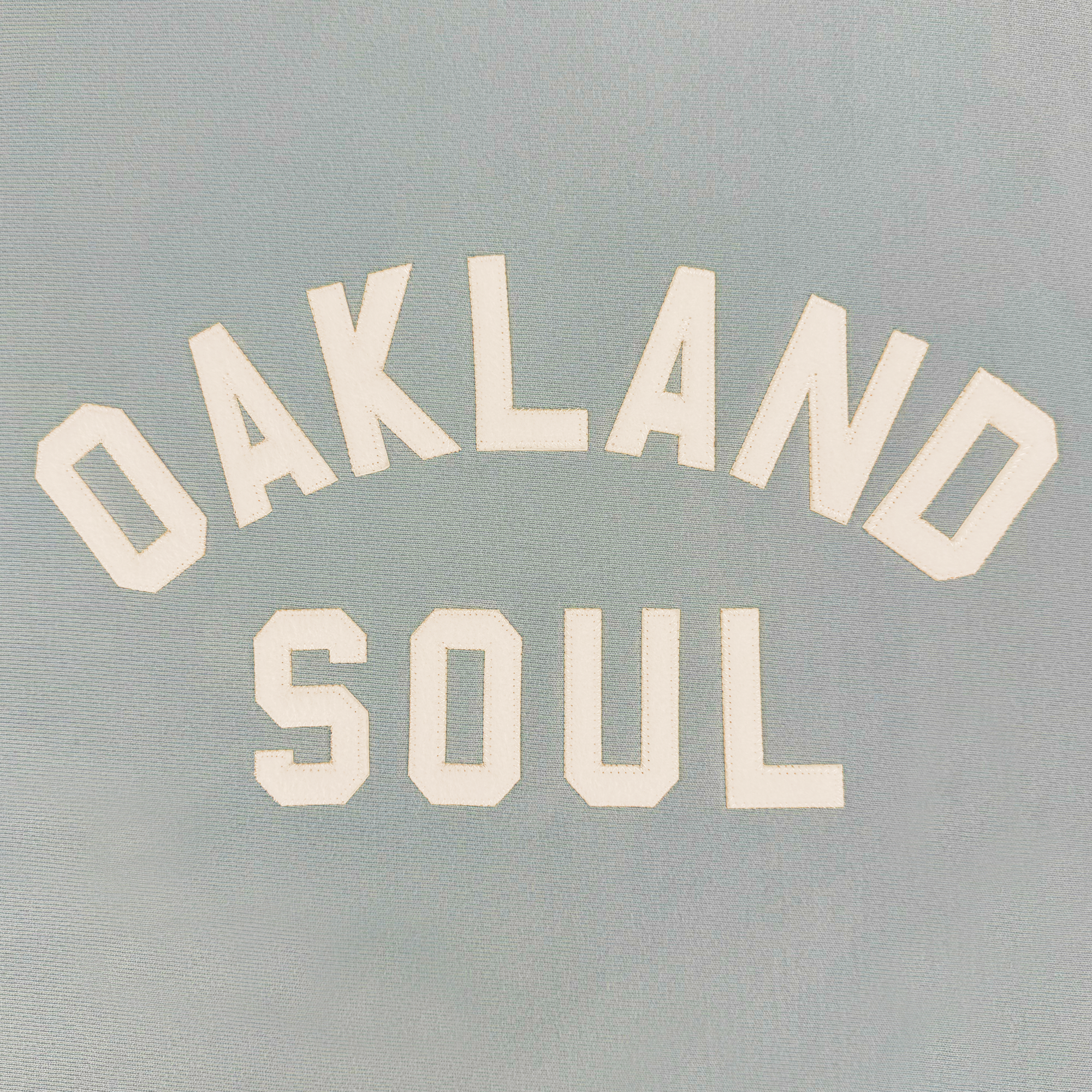 The Champion X Oakland Soul Wordmark Crew displays "OAKLAND SOUL" in bold, white capital letters on a light gray background, arranged in an arch to embody the vibrant spirit of Oakland's women's soccer team and its commitment to social good and community engagement.