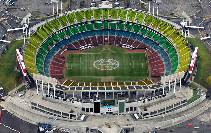 Aerial view of a sports stadium with a predominantly green field marked with "Oakland Roots SC" at its center. The stands are multicolored, featuring green, yellow, red, and blue sections. "Oakland Roots Sports Club" is written prominently at the front of the Oakland Coliseum. Surrounding roads and parking areas are visible. The scene highlights the spirit encapsulated in the Oakland Roots SC Home Tee by Oakland Roots SC.