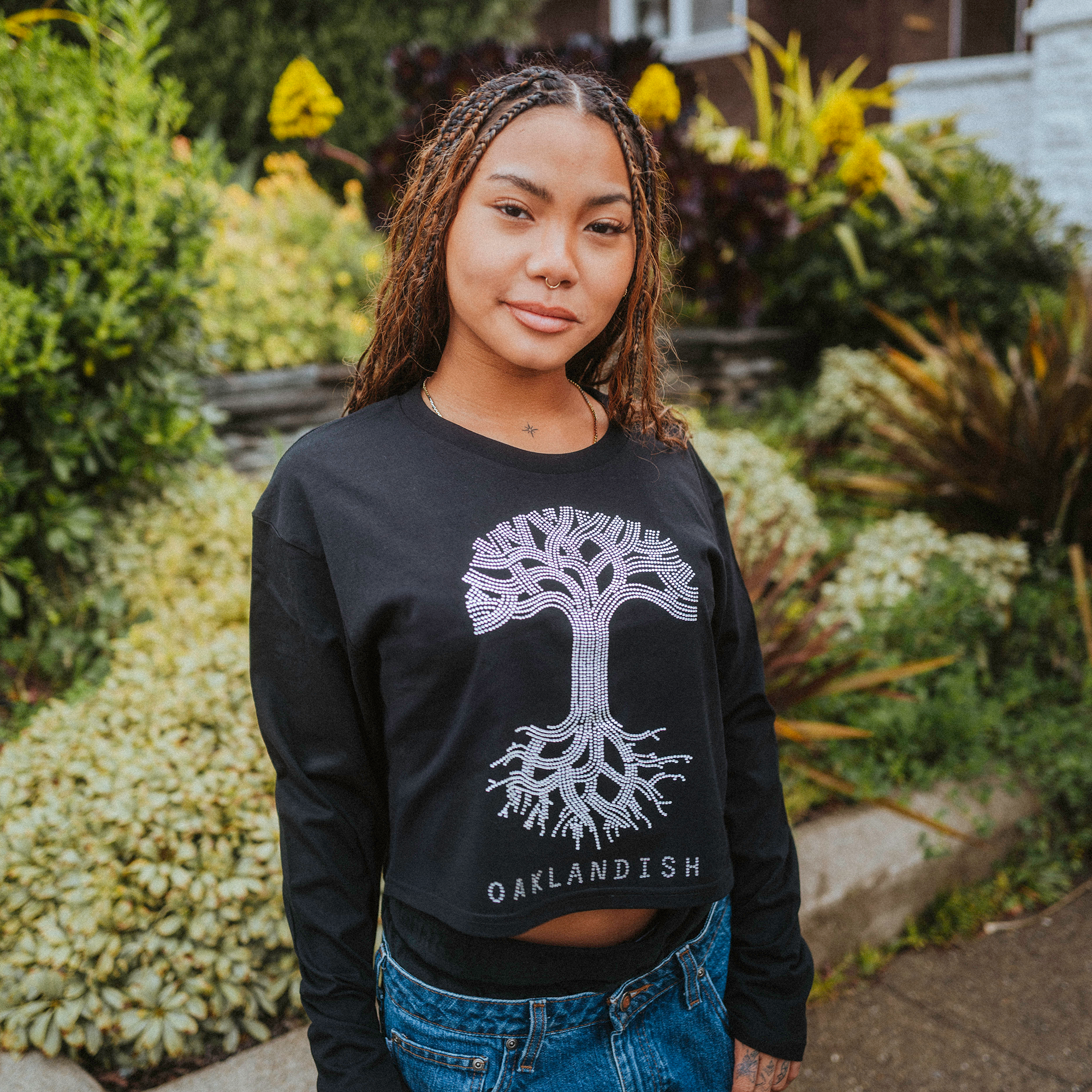 A person with braided hair stands outdoors among green foliage and yellow flowers, wearing blue jeans and a black Women's Oaklandish Classic Logo Rhinestone LS Tee, featuring a rhinestone tree graphic. They confidently look at the camera. Available in women's sizes.