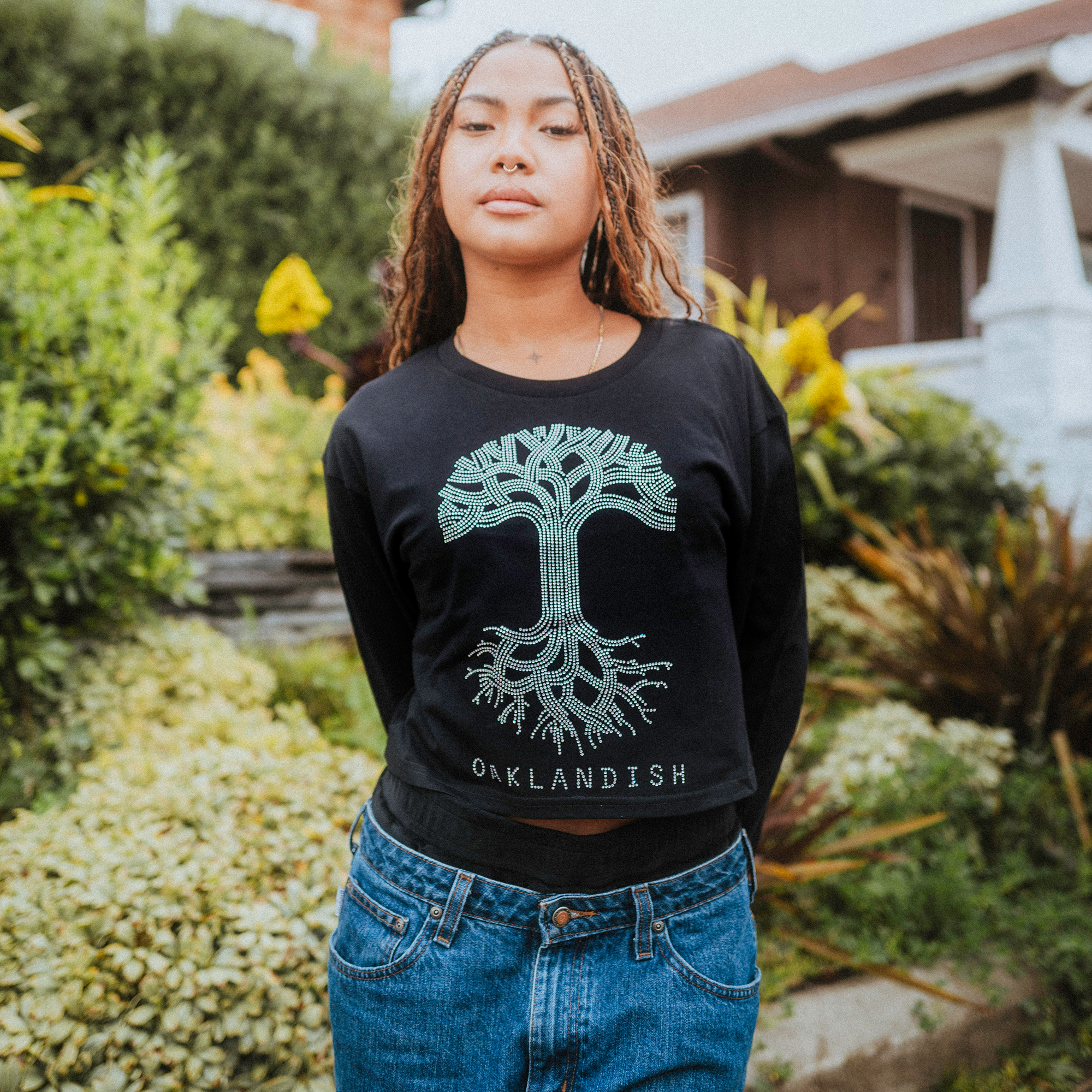 A woman with long braided hair poses outdoors in an Oaklandish Women's Classic Logo Rhinestone LS Tee, a chic black cropped design with a rhinestone tree logo and "OAKLANDISH," paired with blue jeans. She stands amid lush greenery next to a brown house featuring a charming white porch.