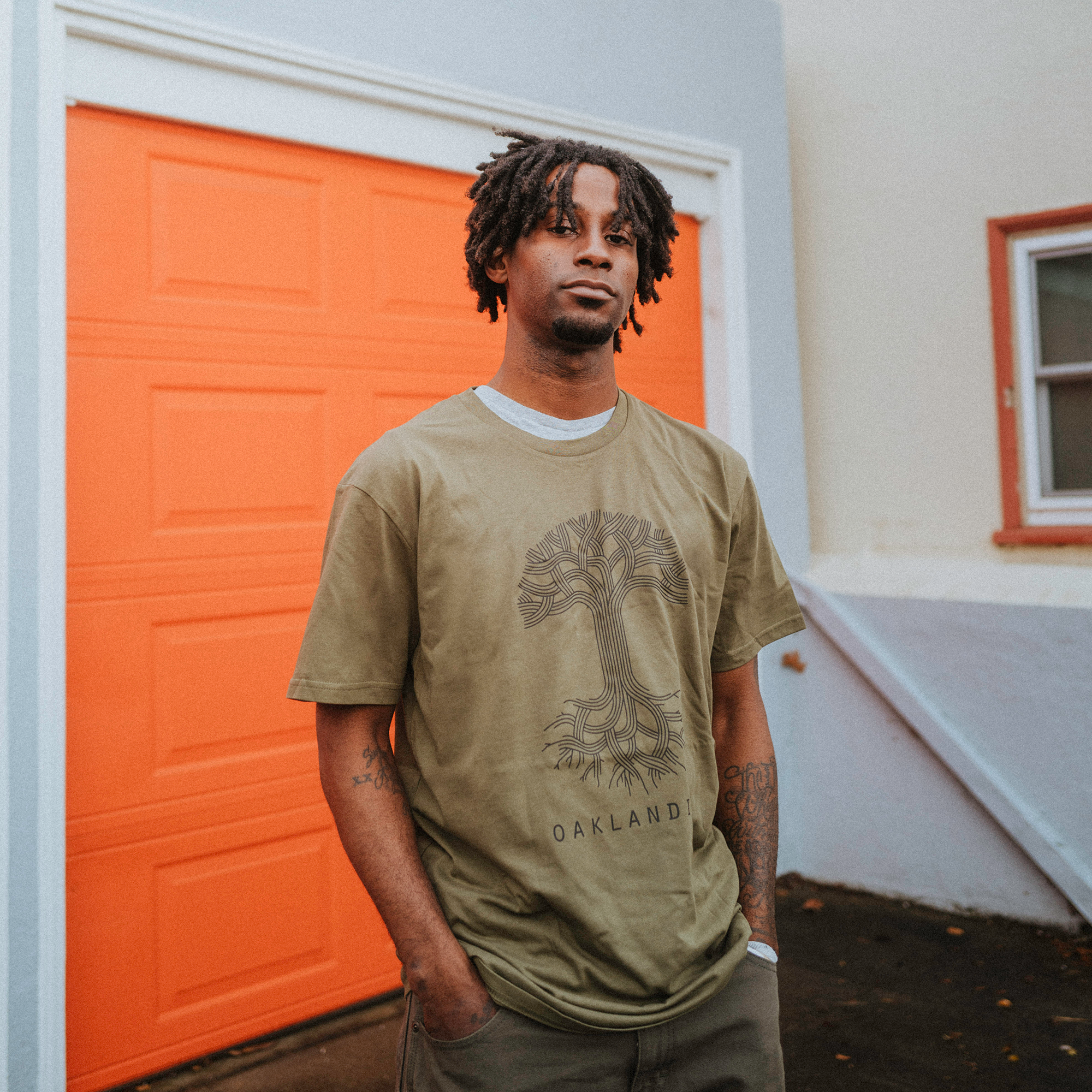 A person with short dreadlocks stands with hands in pockets on a rainy day, wearing the 100% cotton Oaklandish Classic Logo Tee—a green "Oakland" T-shirt with a tree design—capturing the purist style. They are positioned before an orange garage door reflecting rainwater.