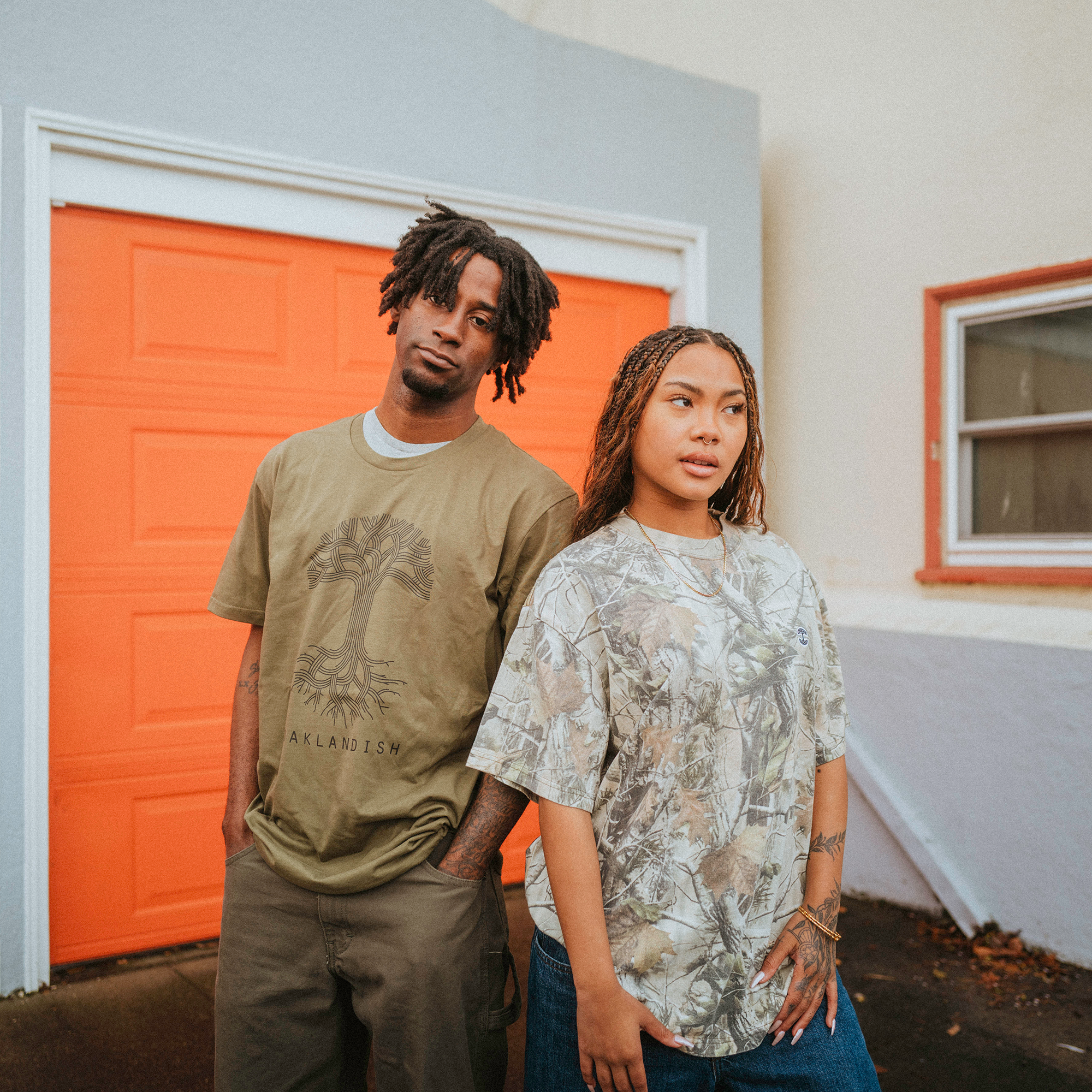 A man wearing a green "AFRICAN LANDISM" heavyweight cotton shirt and a woman in an Oaklandish Camo Tree Tee stand in front of an orange garage door. Both have braided hair and jeans, gazing slightly away from the camera with a relaxed vibe.