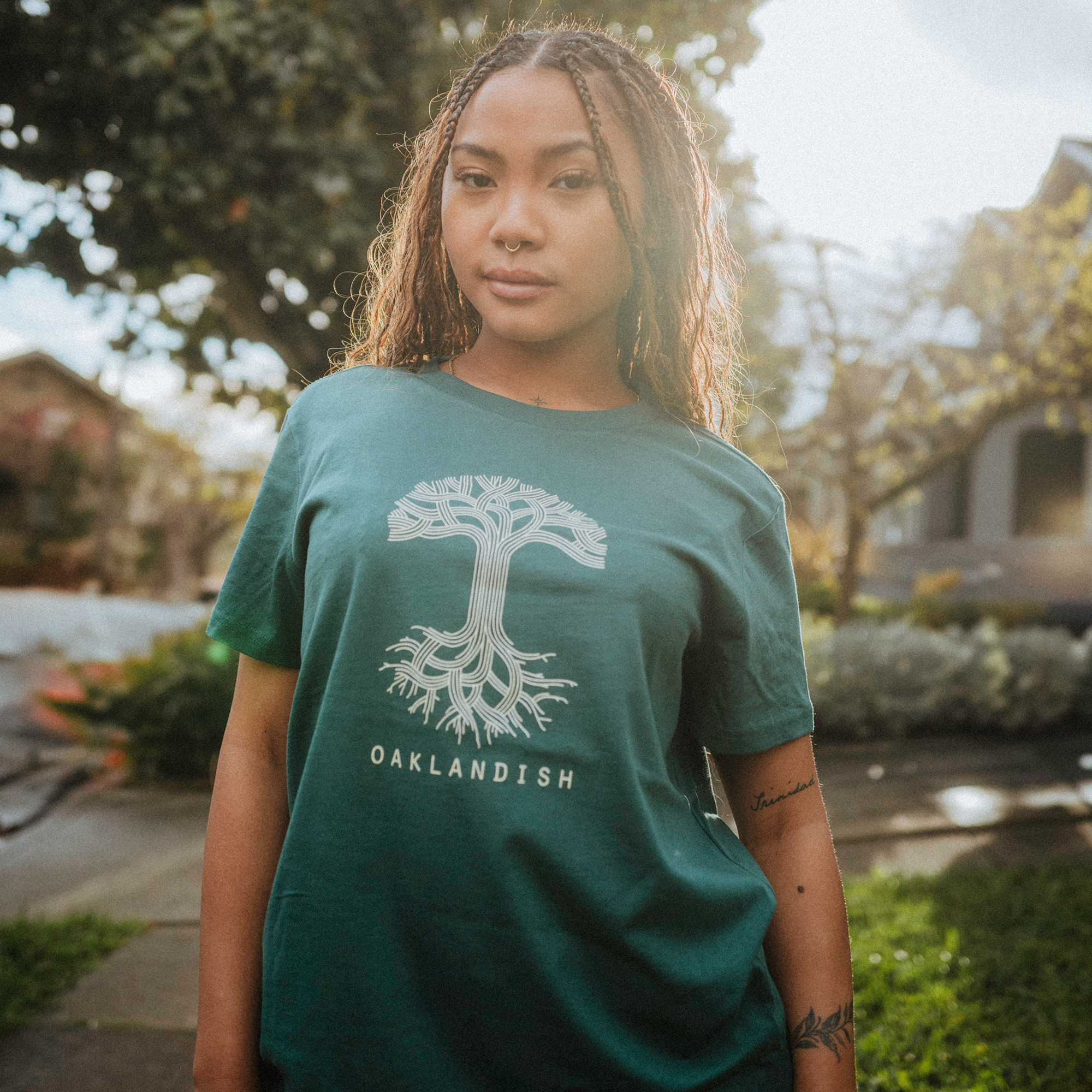 A woman with braided hair stands outdoors wearing the Women's Oaklandish Classic Logo Tee in 100% cotton, featuring a tree design and "Oaklandish." The backdrop of a sunlit tree and house complements her serene demeanor, reminiscent of a tranquil Jade Shop atmosphere.