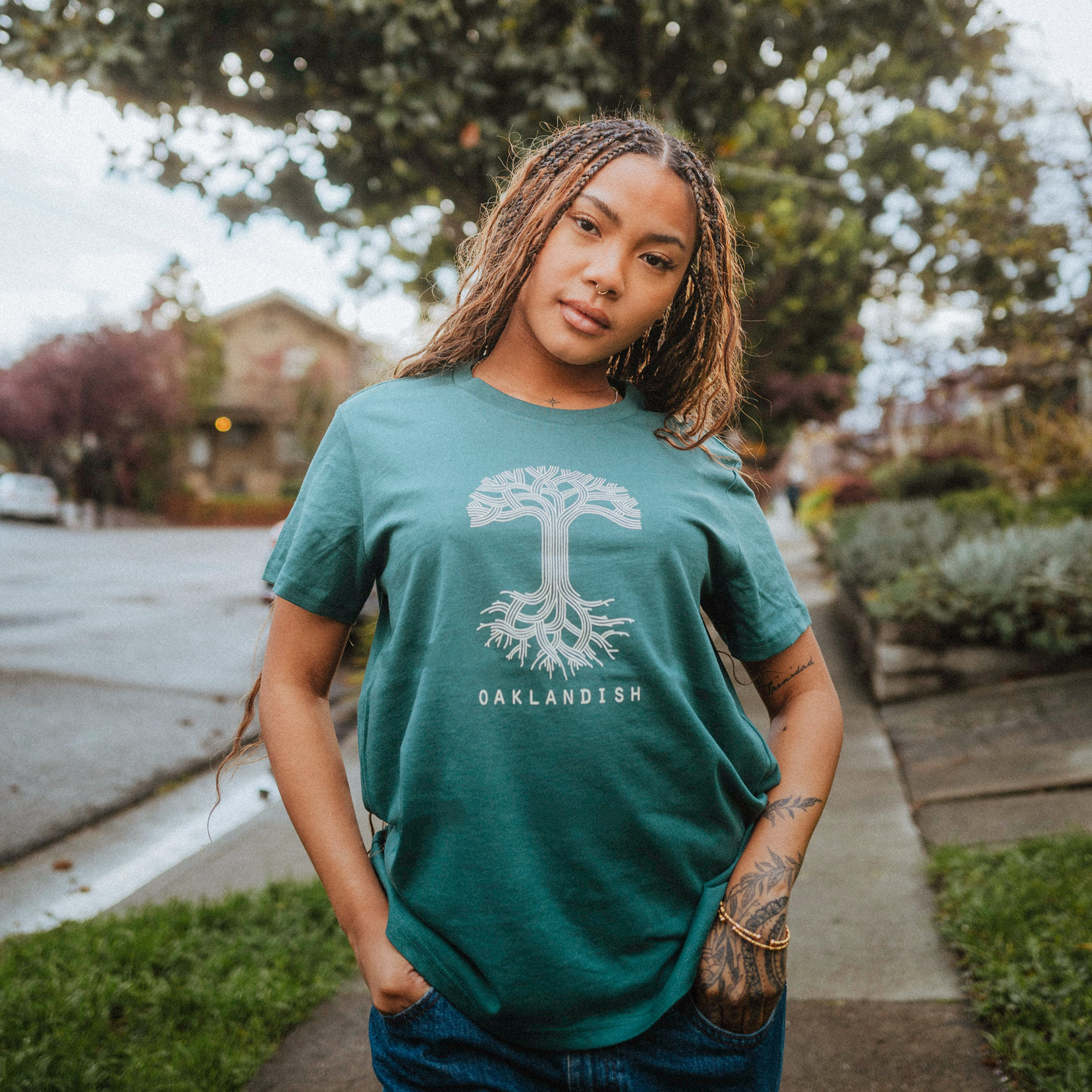 A young woman with braided hair stands on a suburban sidewalk wearing the Women's Oaklandish Classic Logo Tee from Oaklandish. With hands in her jeans pockets, her confident posture is highlighted by her tattooed left forearm against the overcast sky.