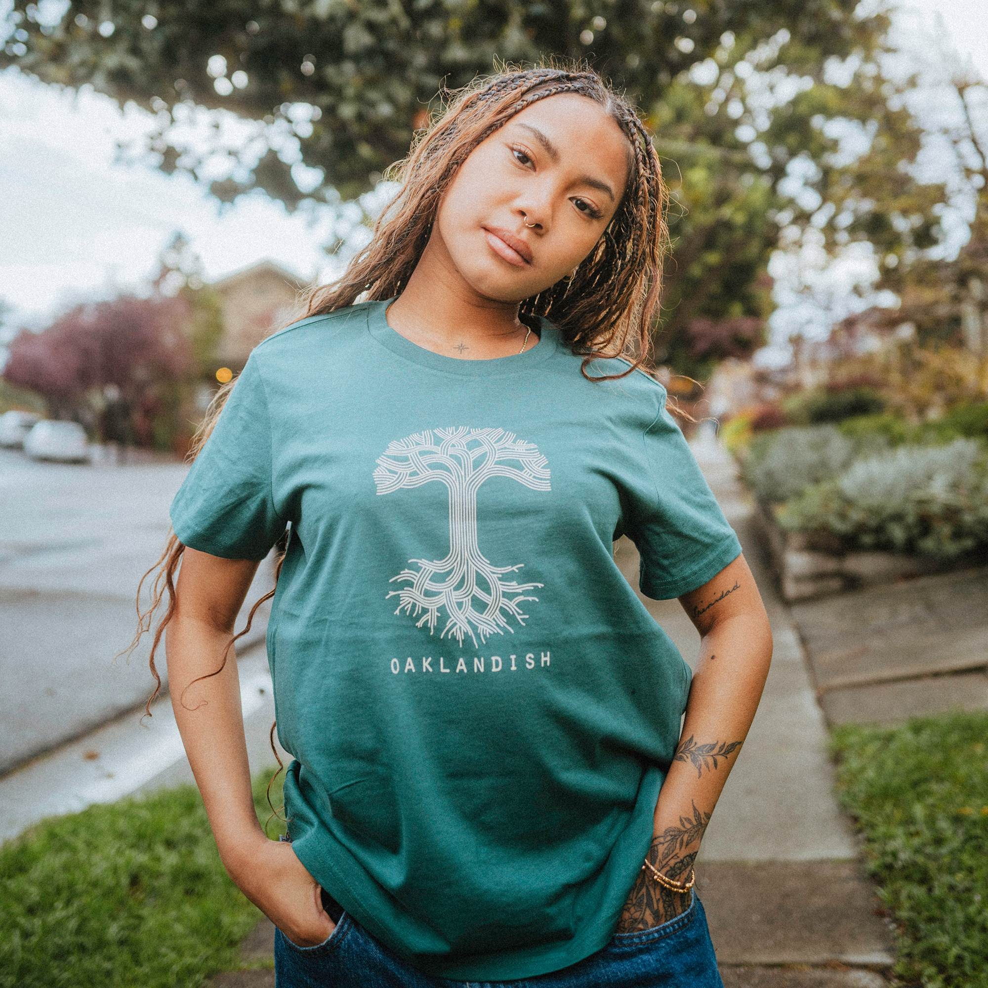 A woman with long, braided hair poses confidently outdoors on a cloudy day, wearing an Oaklandish Women's Classic Logo Tee. The 100% cotton green shirt features a tree design and the text "OAKLANDISH," as she stands on a sidewalk against trees and suburban houses.