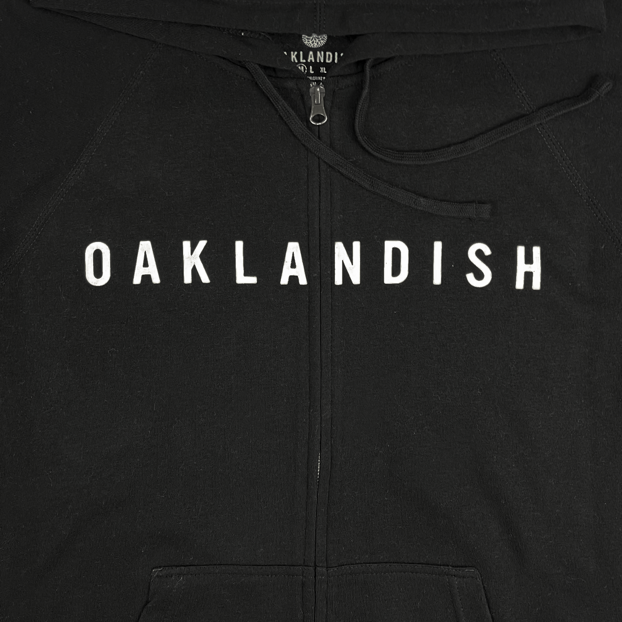 The Oaklandish Classic Zip Hoodie from Oaklandish is a black unisex outerwear item with the word "OAKLANDISH" printed in large, white capital letters across the chest. It features a partially visible front zipper at the top, a drawstring around the hood, and seams near the bottom indicating front pockets. Slim sizing is suggested.