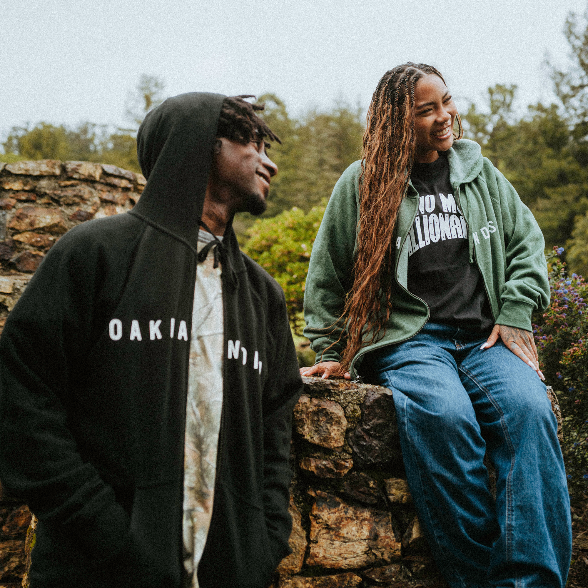 A person in an Oaklandish Classic Zip Hoodie stands by another sitting on a stone wall, smiling. The seated individual, in a green jacket and jeans, enjoys the outdoor scenery with trees behind. Both outfits offer a unisex fit suited for any style.