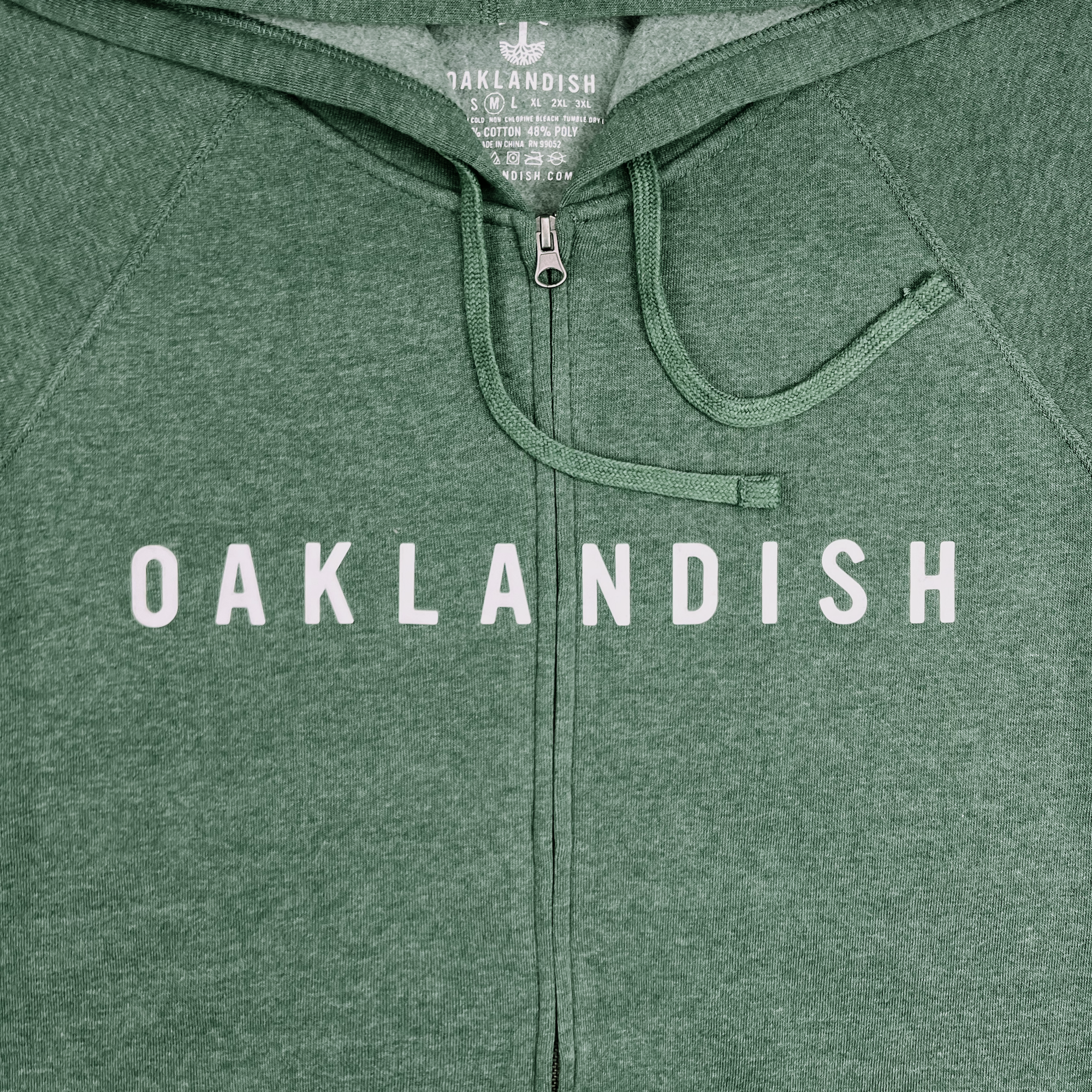 A close-up of the Oaklandish Classic Zip Hoodie in green, featuring "OAKLANDISH" printed in white, uppercase letters across the chest. This unisex outerwear piece from Oaklandish includes an overlapping collar, raglan sleeves, and standard drawstrings. The interior neck label displays the brand's logo and additional product information.