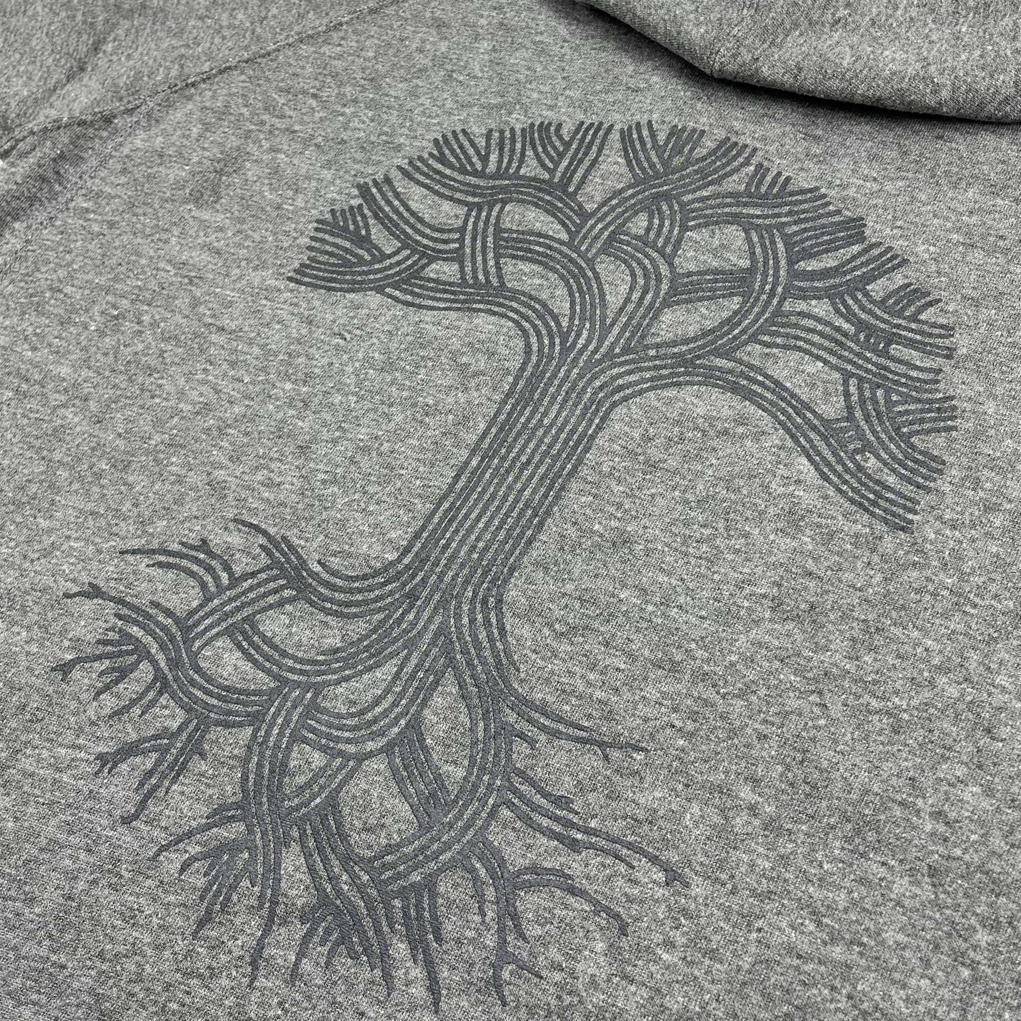 Close-up of the fabric of the Oaklandish Classic Zip Hoodie by Oaklandish, showcasing a stylized black tree design. The design features a thick trunk with intricate, flowing branches and roots extending outward, creating a symmetrical and abstract pattern. This zip-up hoodie appears soft and comfortable, with the tree design adding a unique and artistic touch.