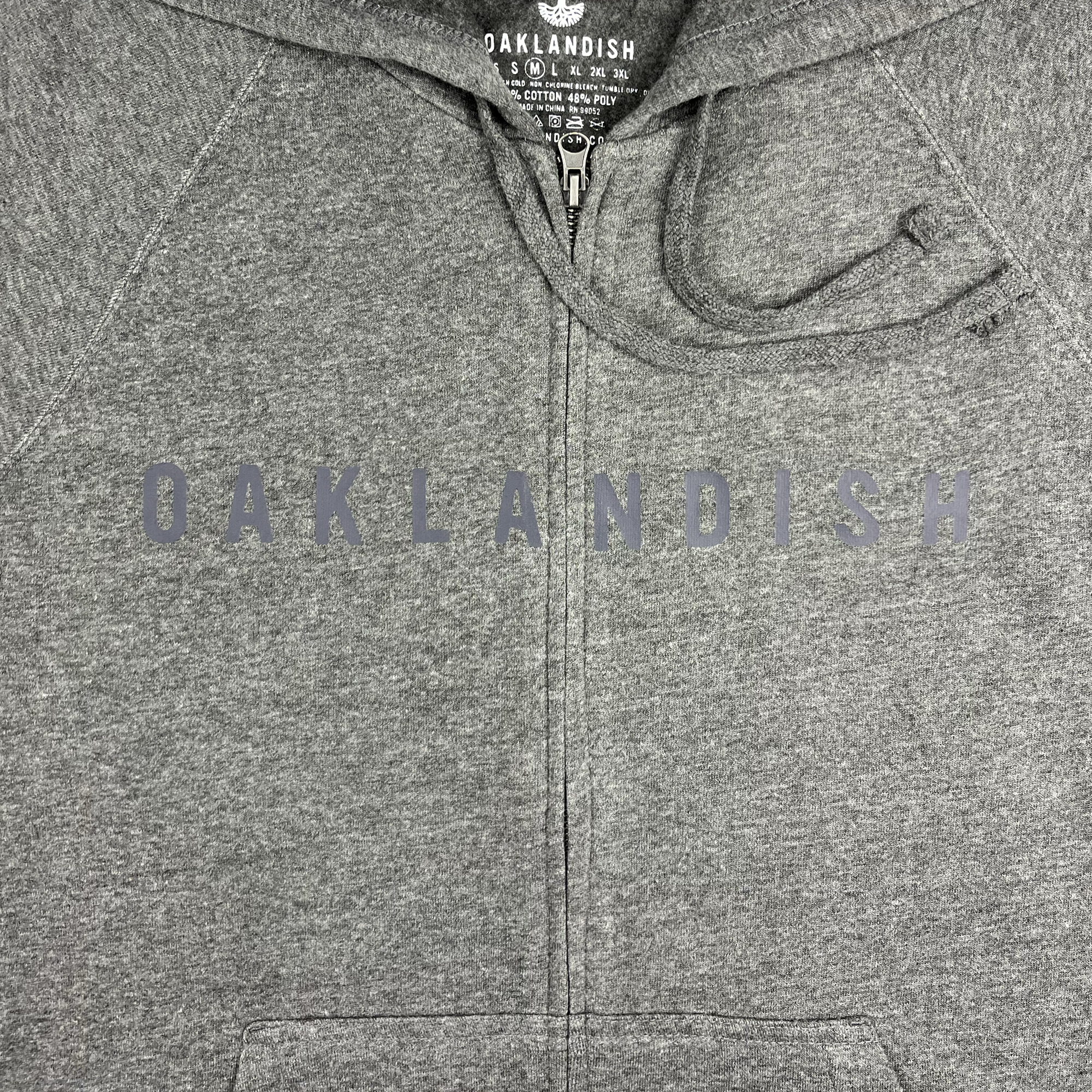 A close-up image of the Oaklandish Classic Zip Hoodie in gray, featuring "OAKLANDISH" prominently printed across the chest in dark, bold letters. This unisex outerwear includes a brand tag inside the collar, displaying the Oaklandish logo, website URL, and hoodie size in white text against a black background.