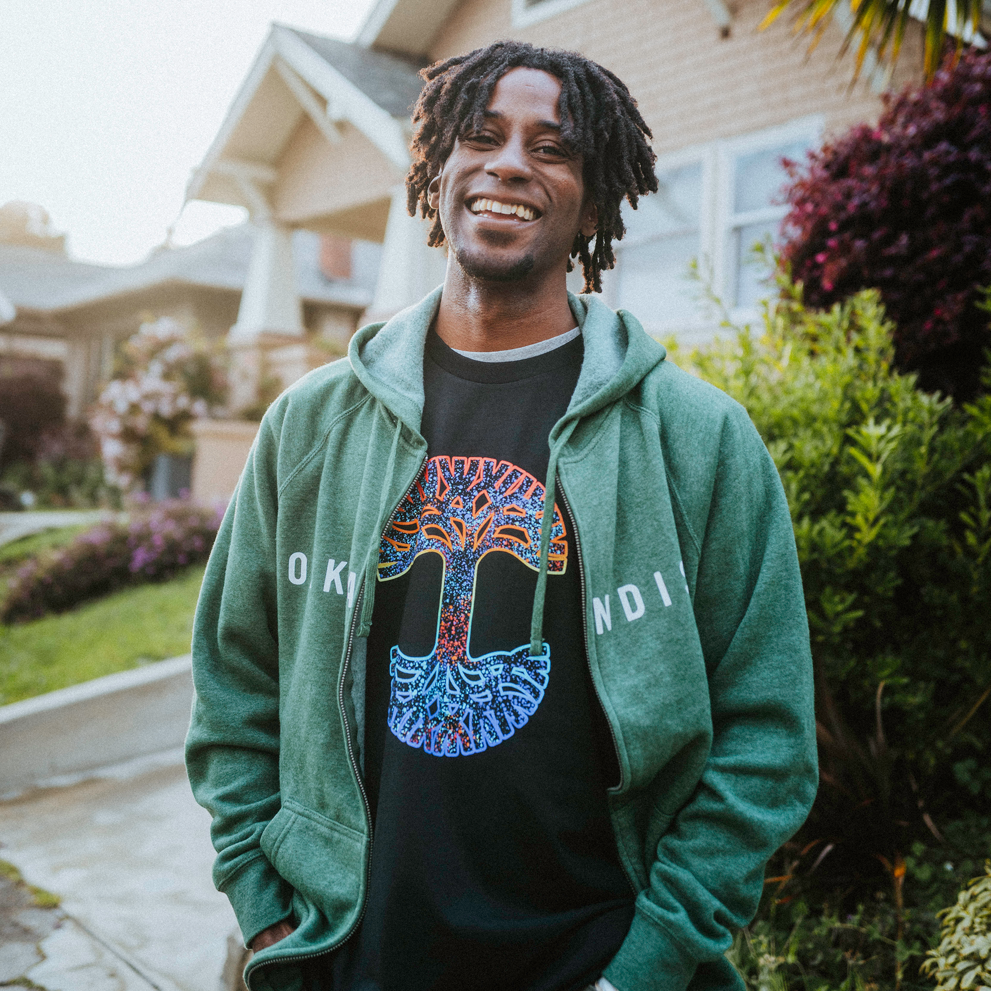 A person with short dreadlocks smiles while standing outdoors, wearing an Oaklandish Classic Zip Hoodie with a comfortable unisex fit over a black T-shirt showcasing a colorful tree design. In the background, houses, lush green plants, and purple flowers line the pathway.