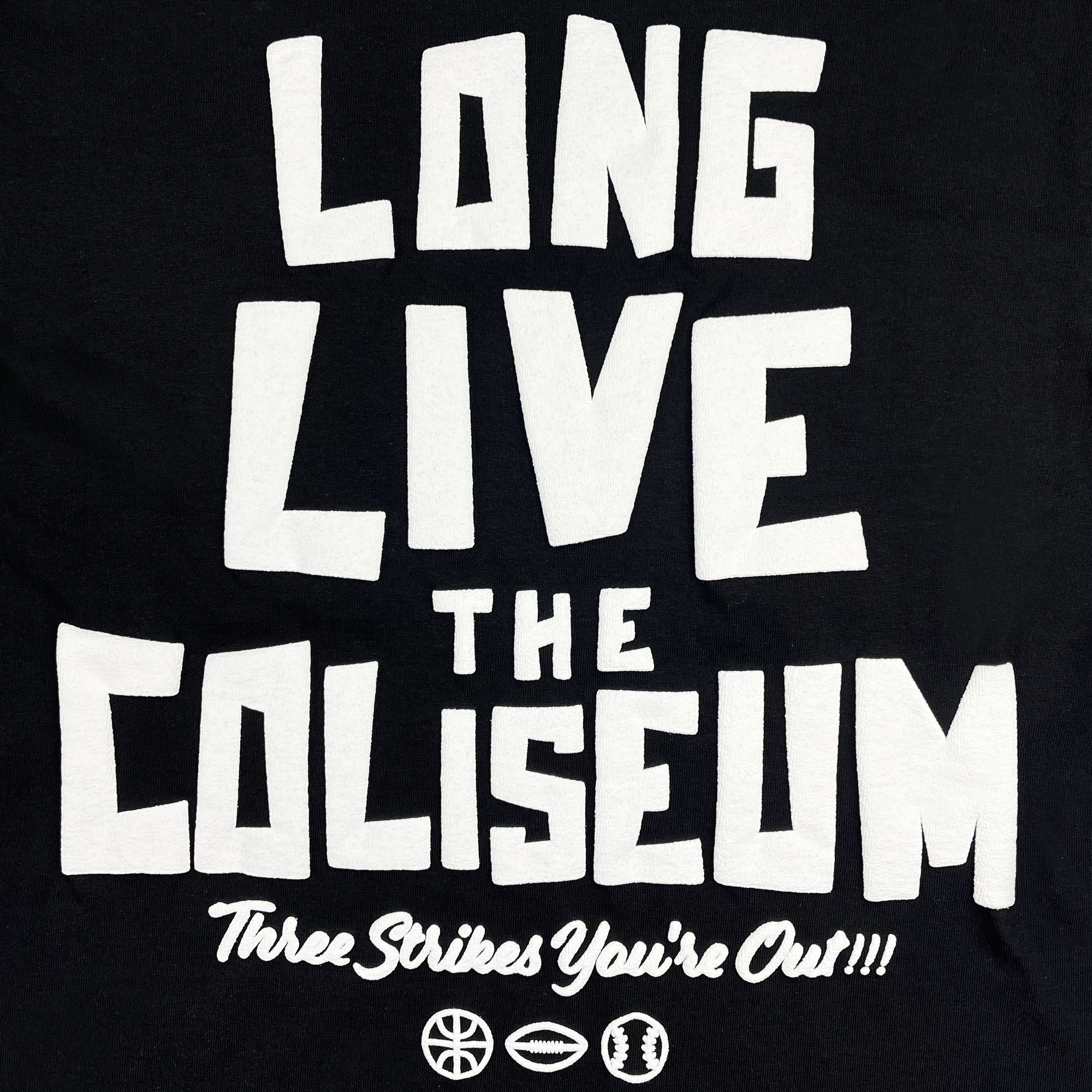 The Coliseum Tee by DOC from Oaklandish is a black t-shirt with bold white text reading, "LONG LIVE THE COLISEUM" in large block letters. Below that, it says, "Three Strikes You're Out!!!" in smaller text. Designed by Dustin O. Canalin, the tee features four small sports icons—a basketball, a soccer ball, a football, and a baseball—at the bottom.