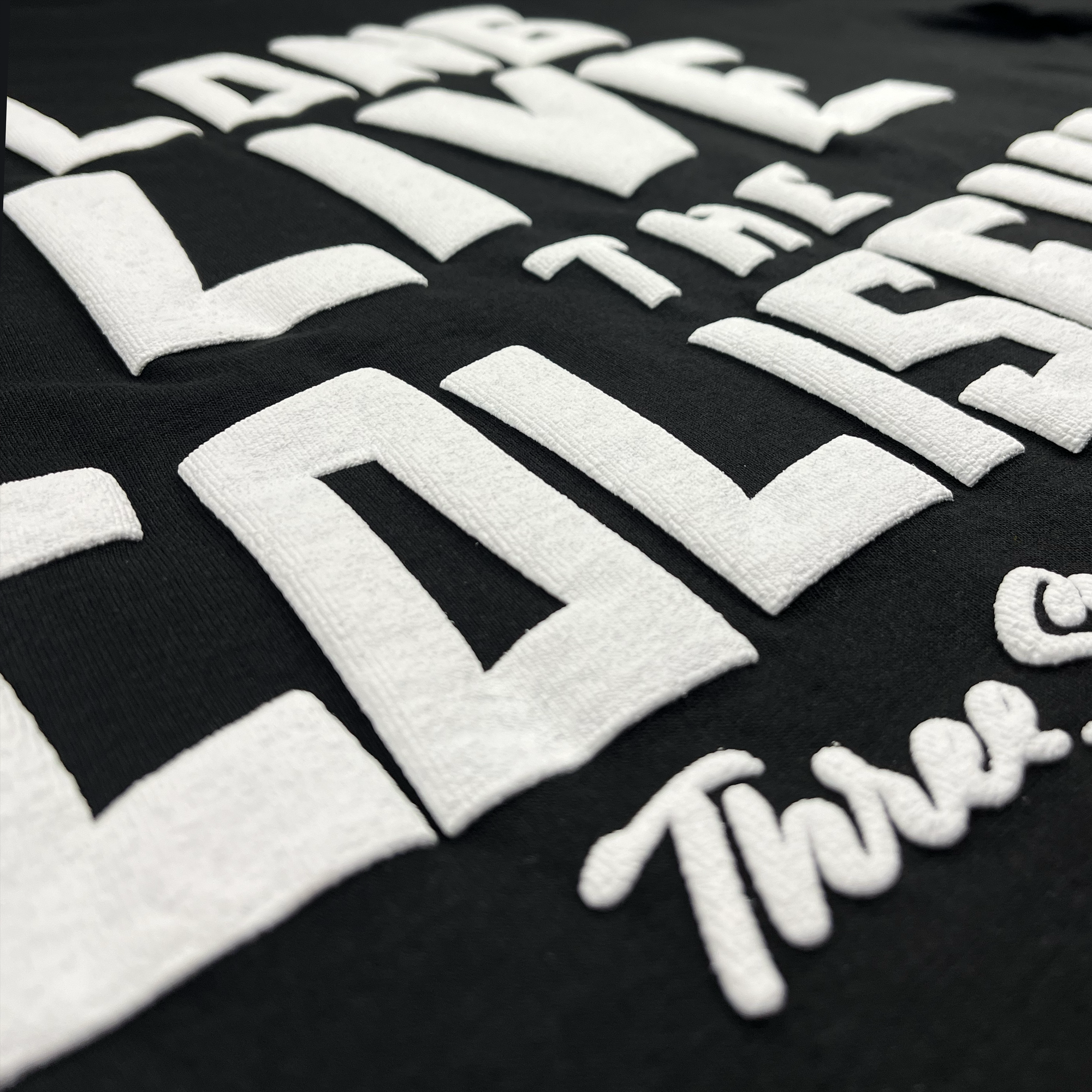 Close-up view of a black fabric with raised, thick white text that reads, "Long Live the Coliseum Three-Sixty." The words are bold and capitalized, with some smaller cursive writing at the bottom right. This Oaklandish Coliseum Tee By DOC is perfect final game memorabilia for any loyal Oakland pro baseball fan.