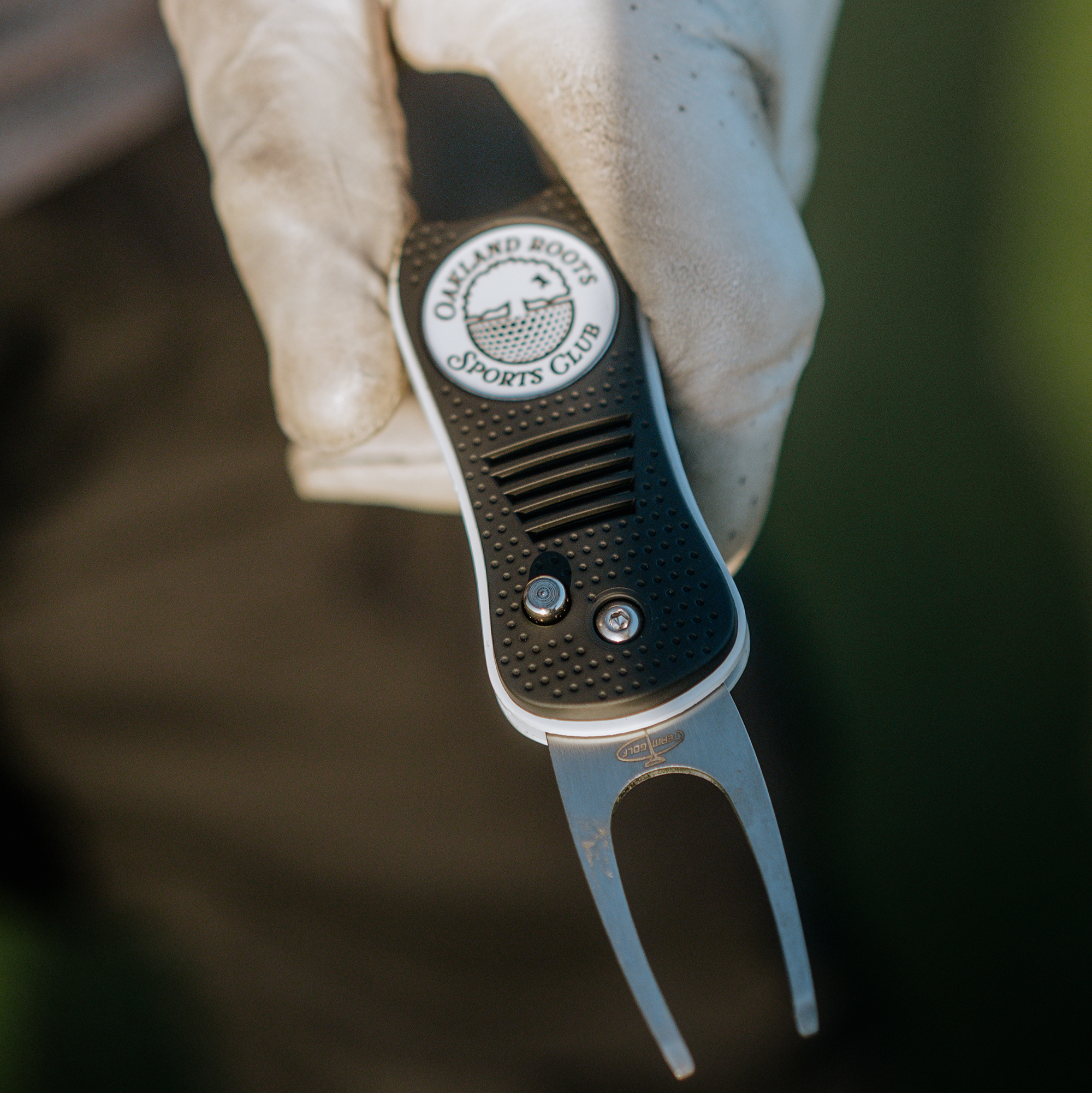 A person wearing a white glove holds the Heritage Divot Tool, a black and silver golf accessory by Oakland Roots SC, featuring their distinctive logo of a half soccer ball and tree. The blurred background emphasizes this sleek Oaklandish product.