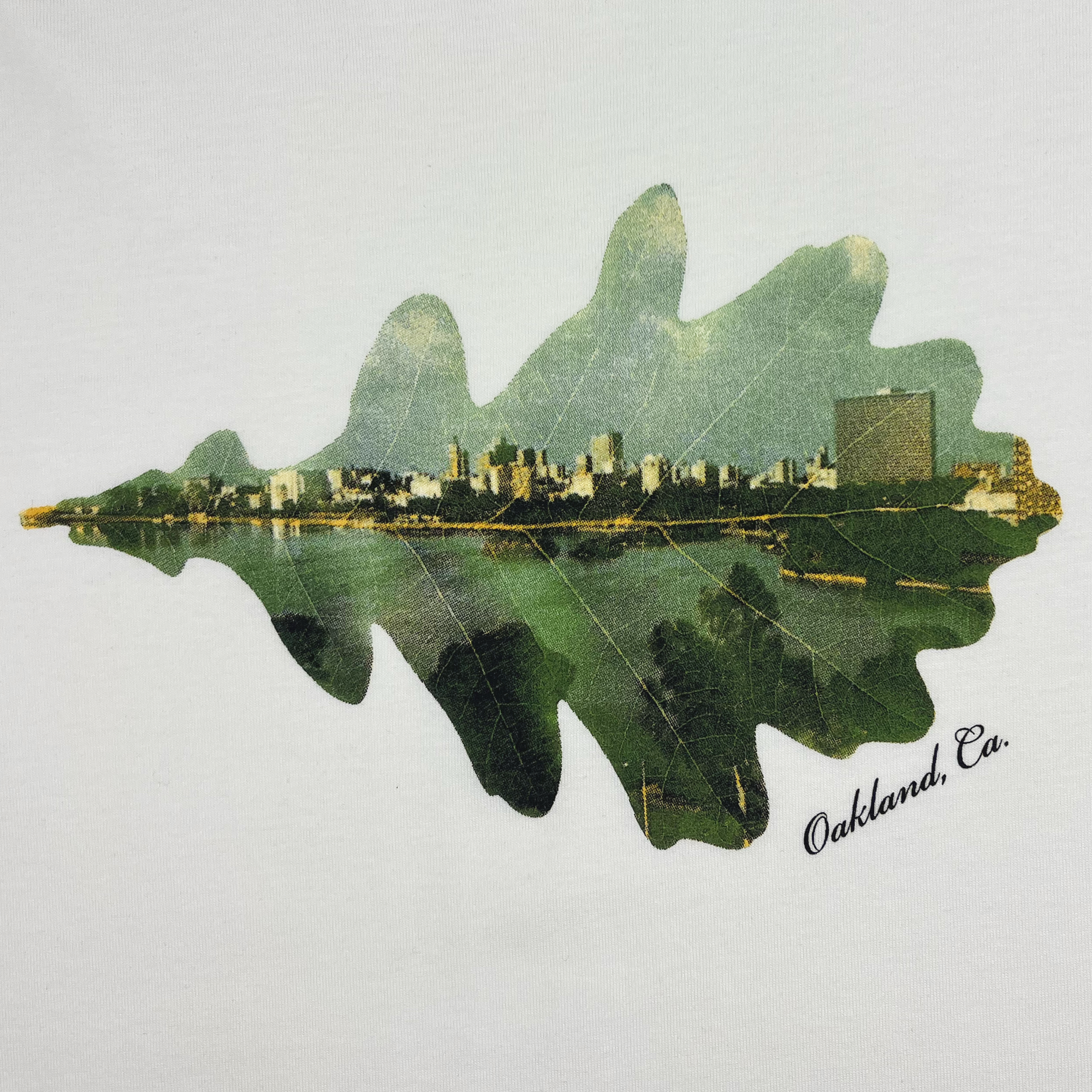 The Women's Double Exposure Tee by Oaklandish features an artistic rendition of the Oakland, California skyline creatively integrated within the outline of a green oak leaf, showcasing the city's natural beauty. Below the leaf, "Oakland, Ca." is elegantly inscribed in cursive on a white background of this women's cotton shirt.