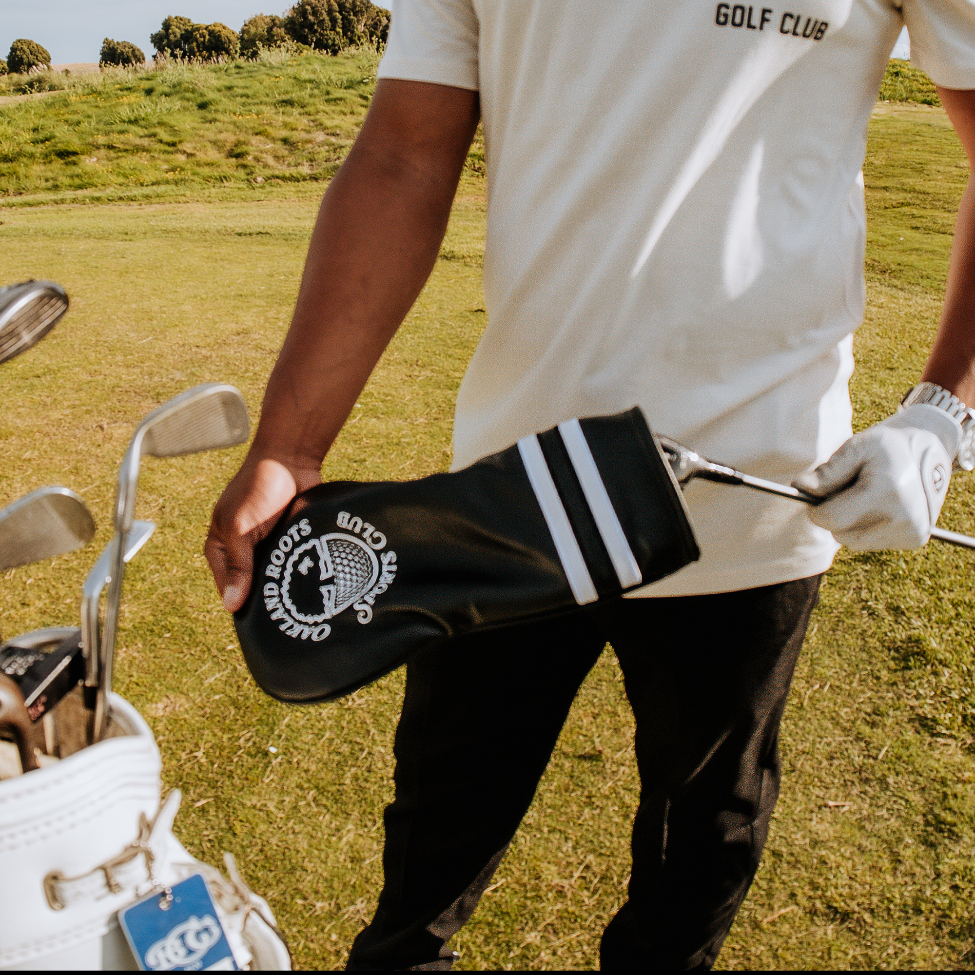 A person stands on a grassy golf course, dressed in black pants and a white shirt that says "GOLF CLUB." In their left hand, they hold a golf club adorned with an Oakland Roots SC Heritage Driver Headcover. A white golf bag filled with clubs is visible in the foreground, embodying the spirit of their sports club.