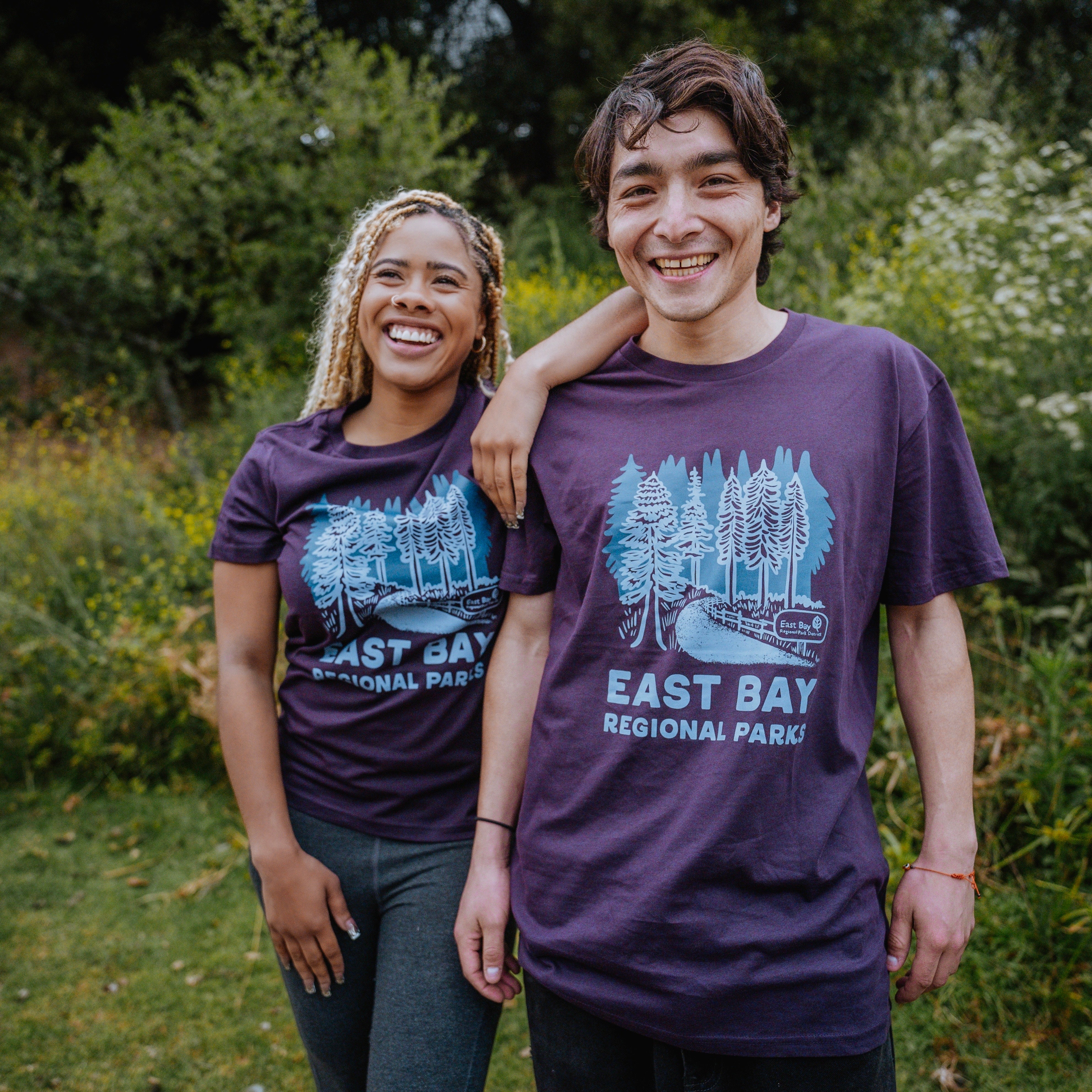 A young woman and man stand outside on grass, both smiling and wearing matching purple "East Bay Regional Parks Tee" classic fit t-shirts by Oaklandish, featuring a graphic of trees. The woman has her arm around the man's shoulders. They are surrounded by lush, green vegetation and wildflowers in the background.